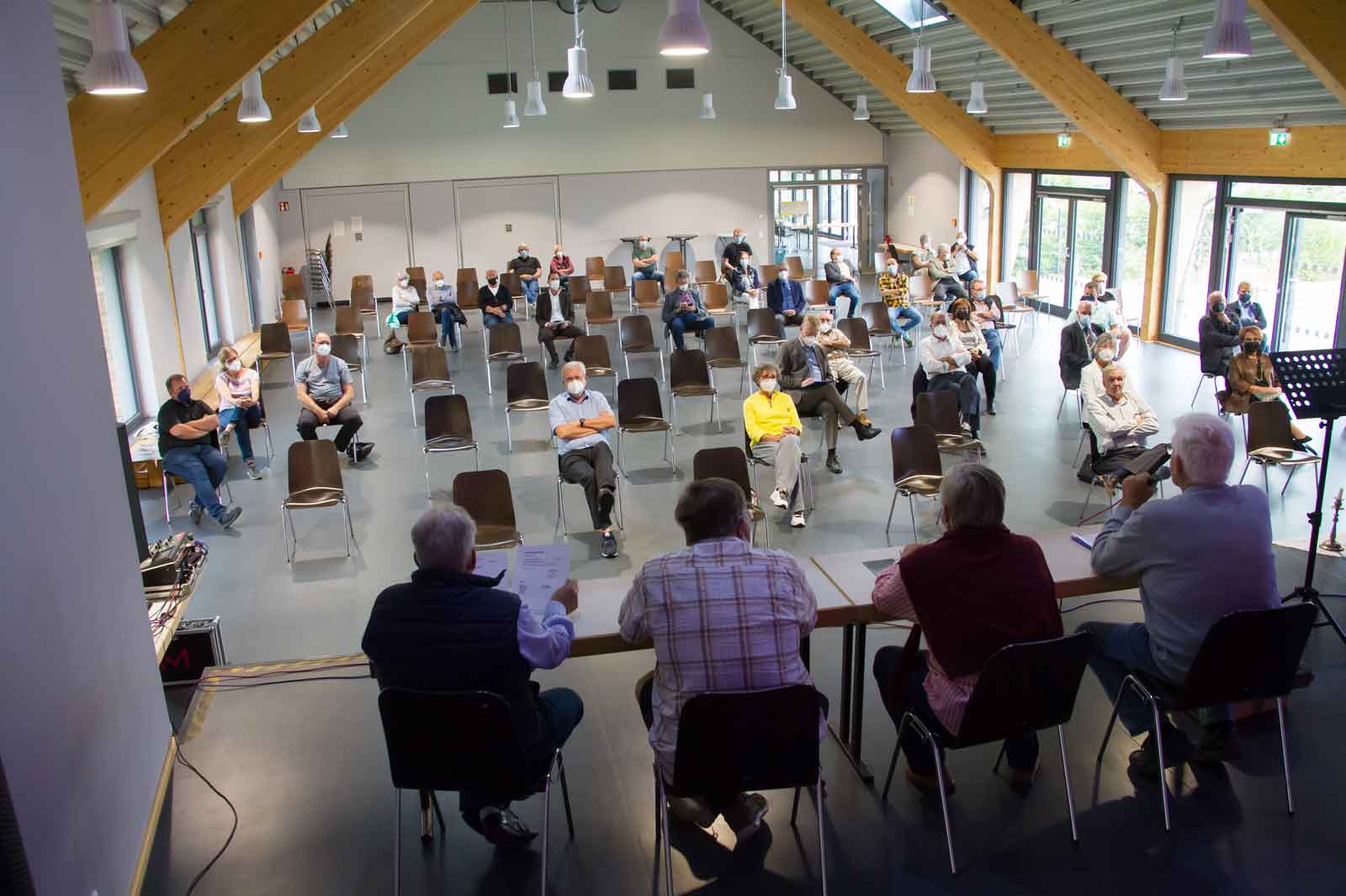 Eine große Versammlung in einem geräumigen Saal mit Stühlen, die für soziale Distanzierung angeordnet sind. Ein Podium spricht zur Menge.