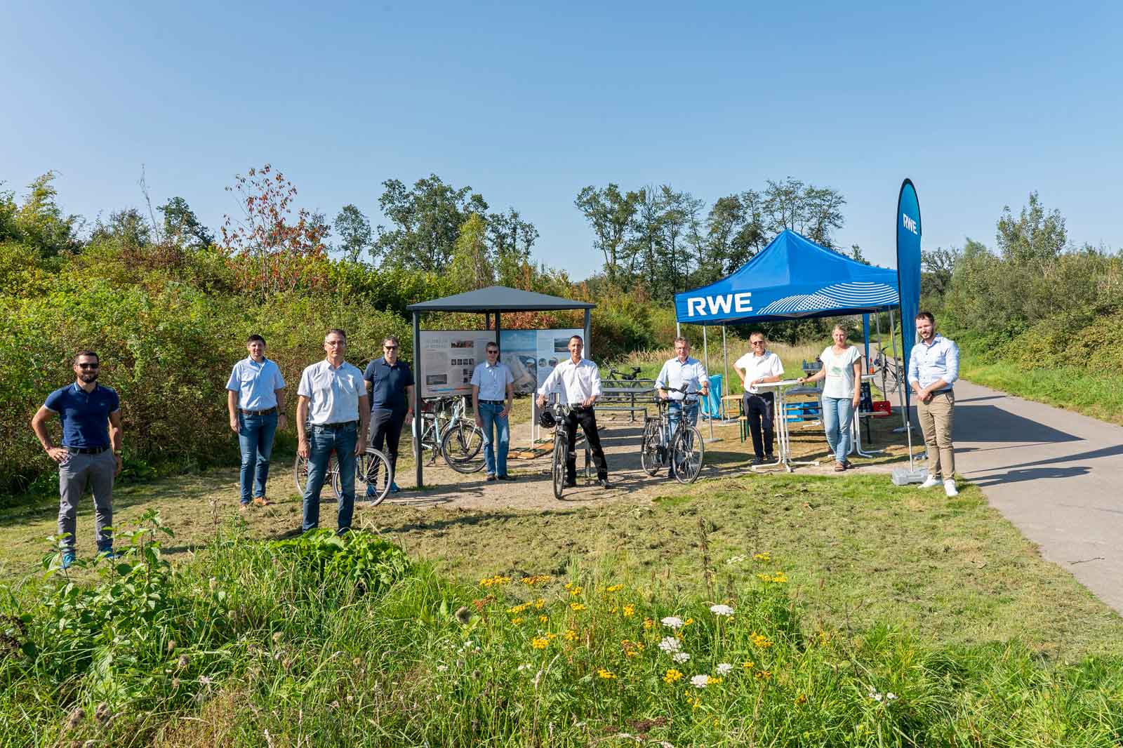 Eine Gruppe von Menschen steht in der Nähe eines blauen RWE Zeltes, mit Fahrrädern und Grünflächen im Hintergrund an einem sonnigen Tag.