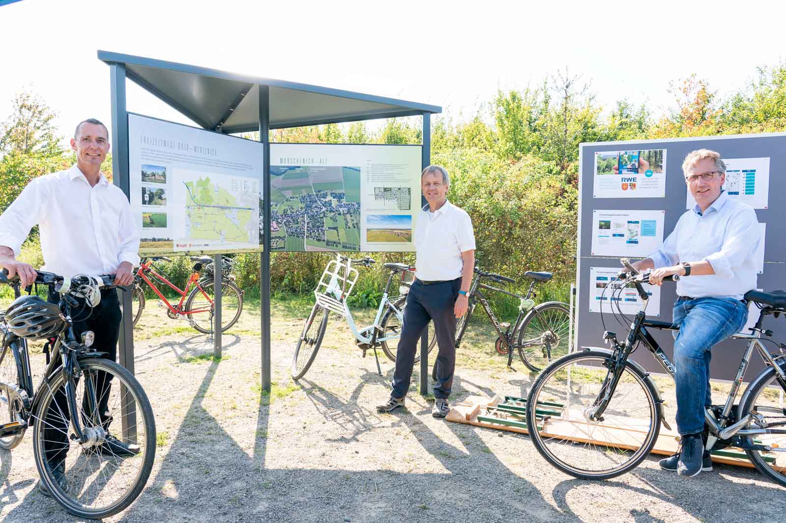 Drei Männer mit Fahrrädern stehen vor Informationsschildern in einer sonnigen Umgebung.