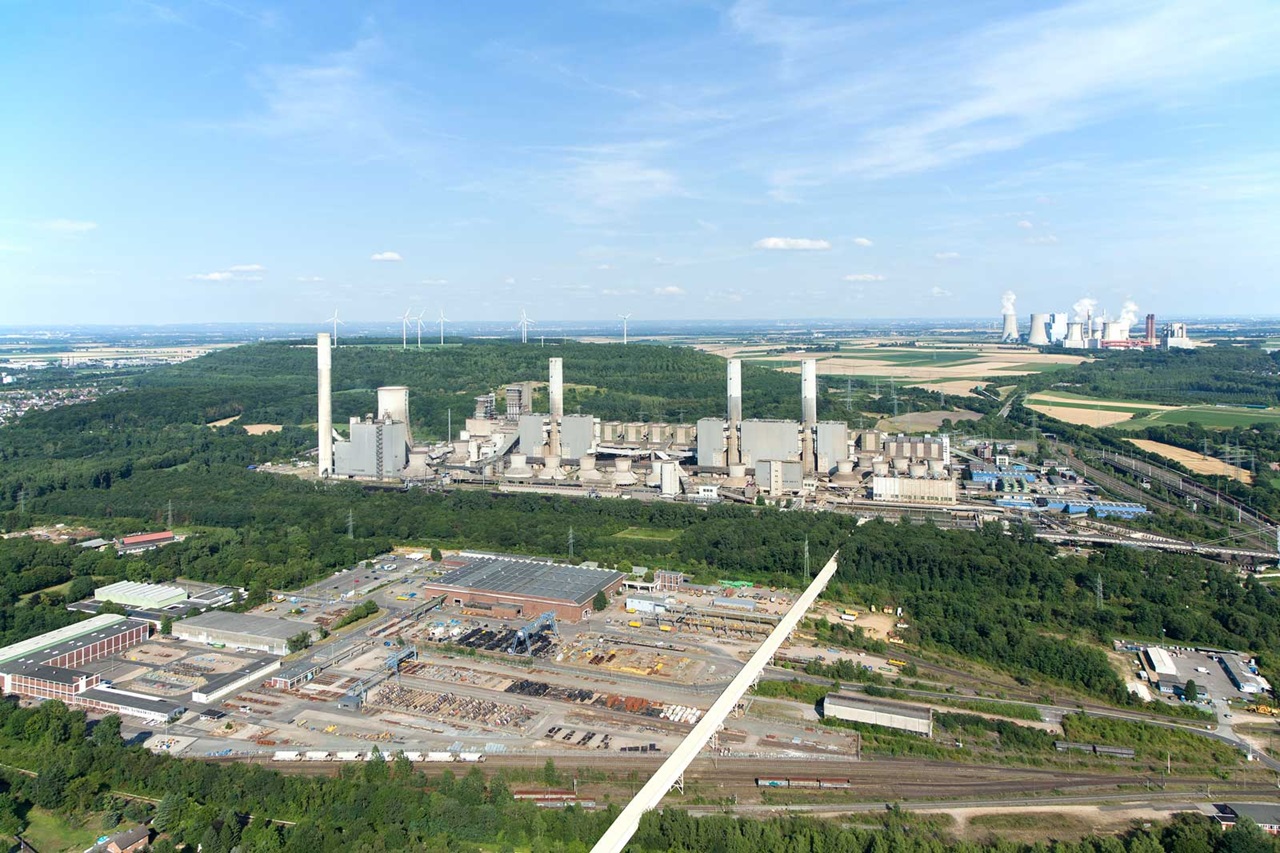 Luftaufnahme eines Industriegebiets mit Kraftwerken, Windturbinen und grünen Wäldern unter einem klaren blauen Himmel.