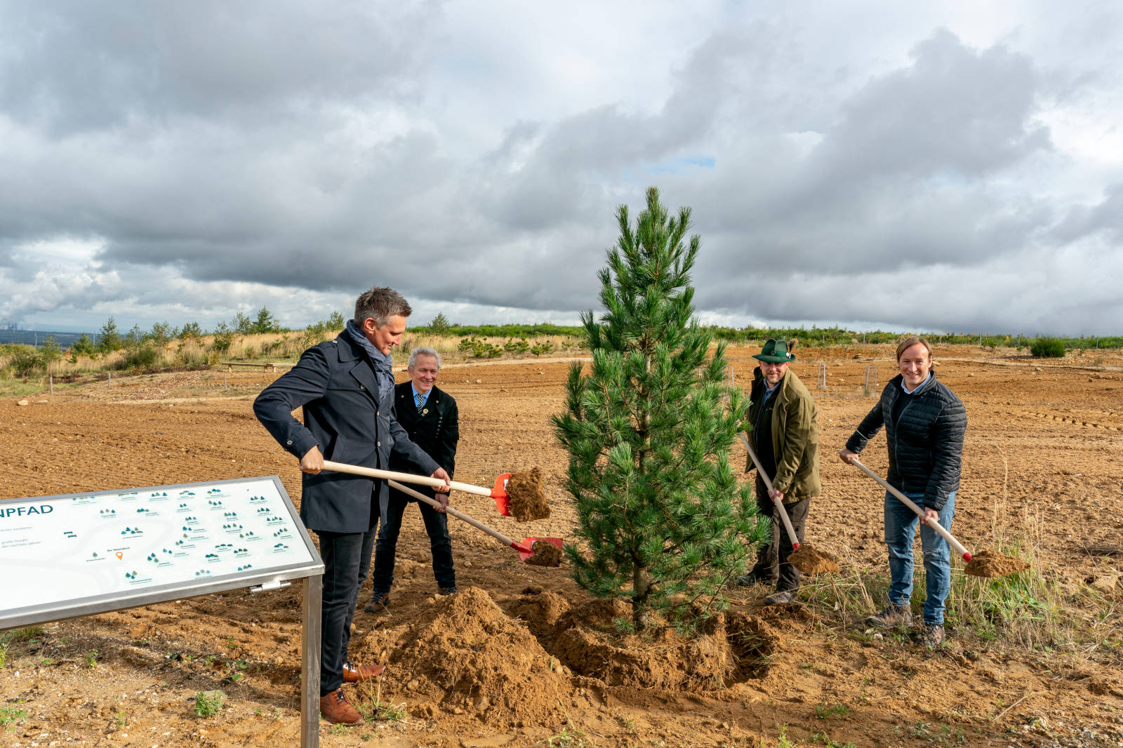 Vier Personen pflanzen einen jungen Baum auf einem Feld, während sie Schaufeln benutzen und von einem klaren Himmel umgeben sind.
