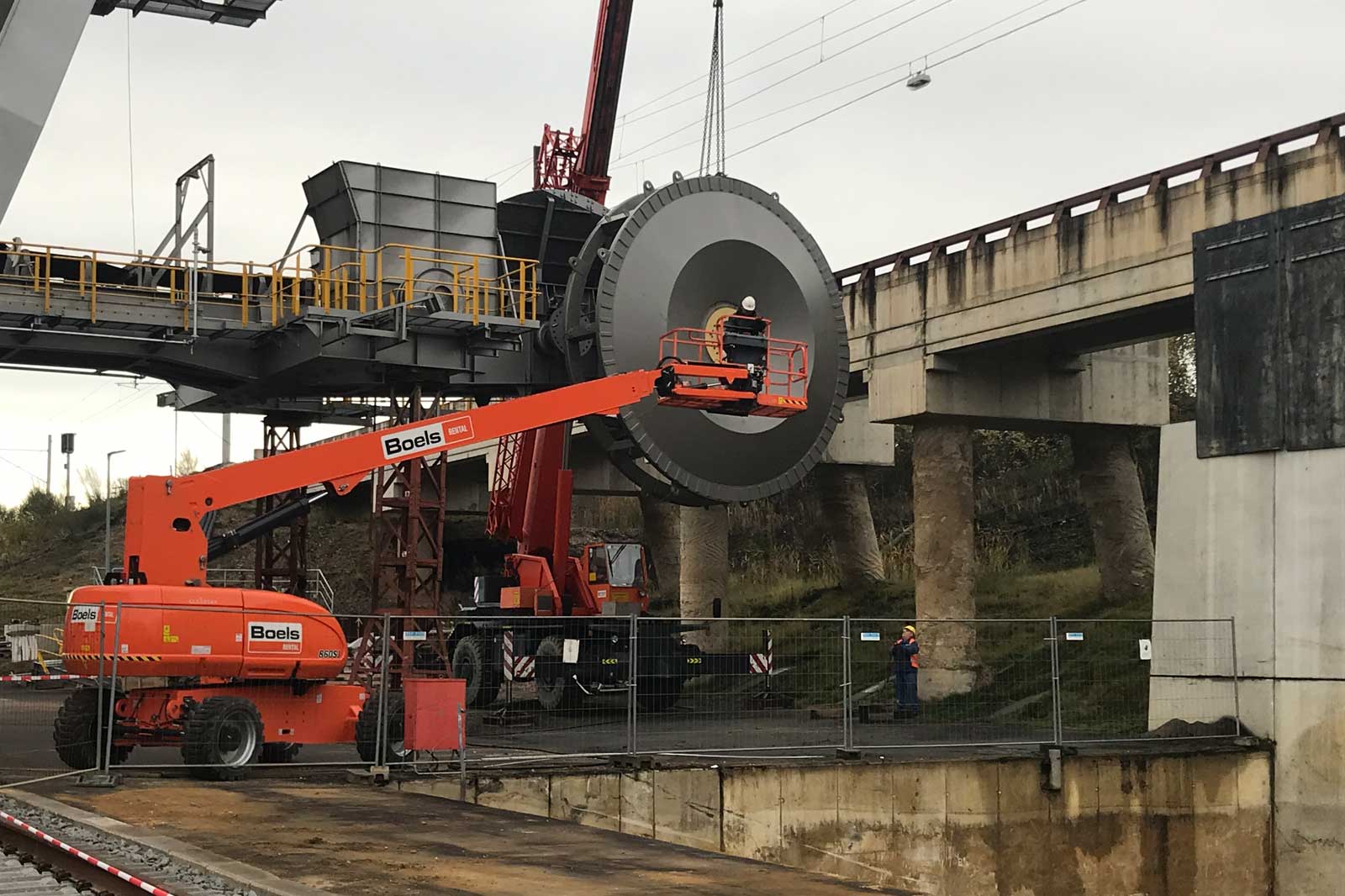 Ein großes Maschinenbauteil wird von einem Kran angehoben, während Arbeiter die Operation überwachen. Sicherheitsbarrieren sind vorhanden.