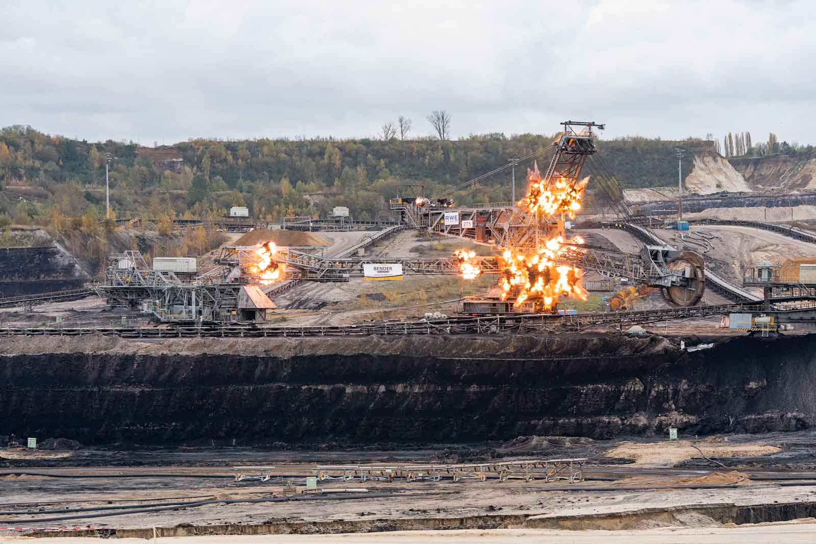 Eine Bergbaustelle mit Maschinen, umgeben von Explosionen und einer rauchigen Landschaft unter einem bewölkten Himmel.