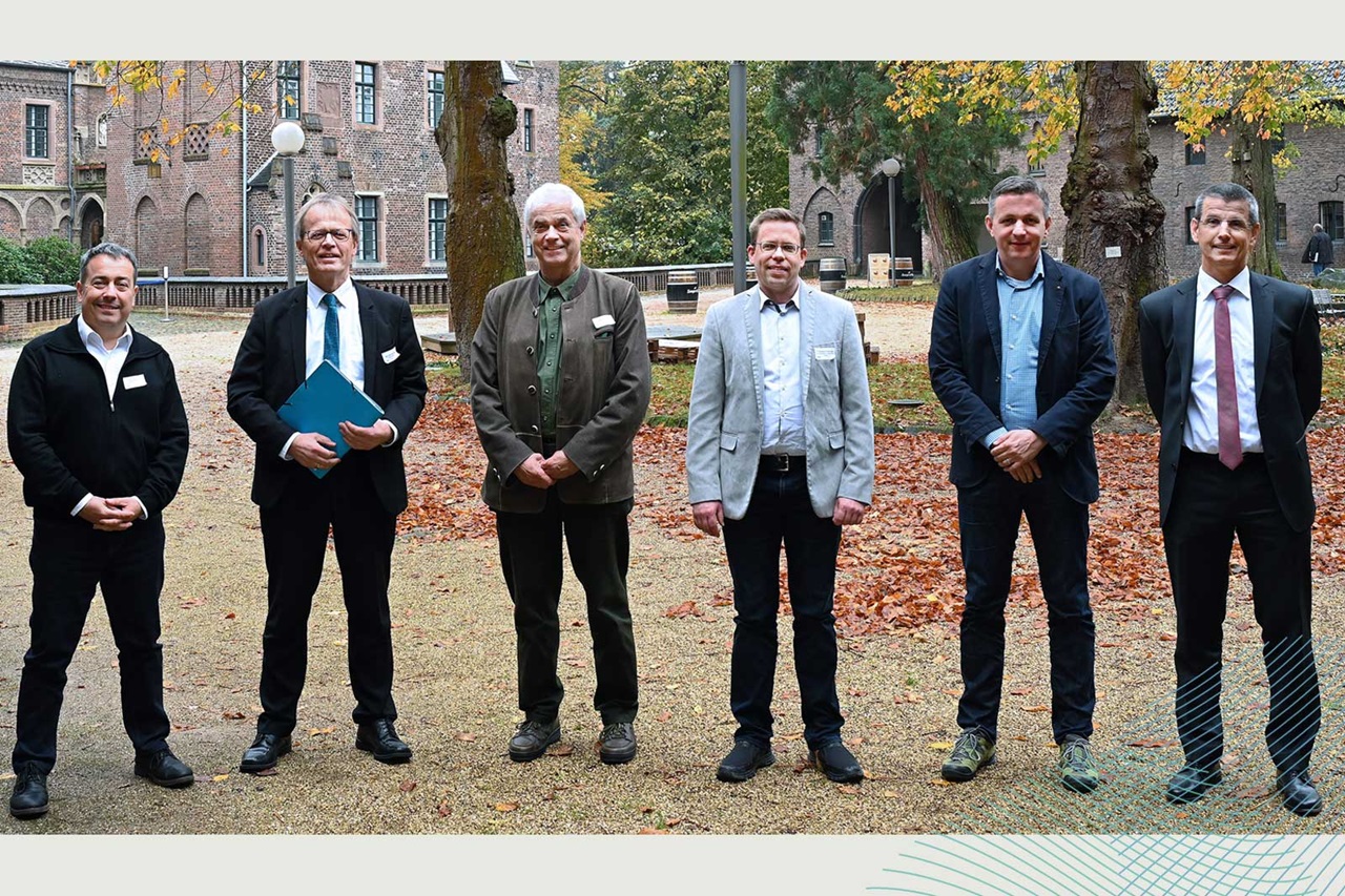 Eine Gruppe von sechs Männern, die formell gekleidet sind, steht im Freien zwischen Herbstblättern in der Nähe eines historischen Gebäudes.