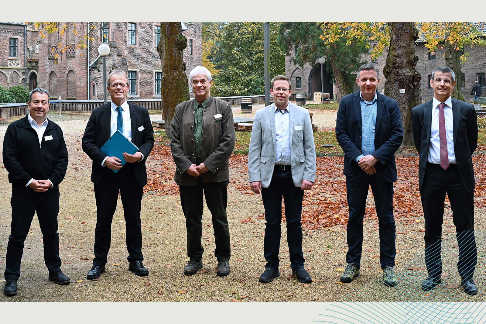 Eine Gruppe von sechs Männern, die formell gekleidet sind, steht im Freien zwischen Herbstblättern in der Nähe eines historischen Gebäudes.