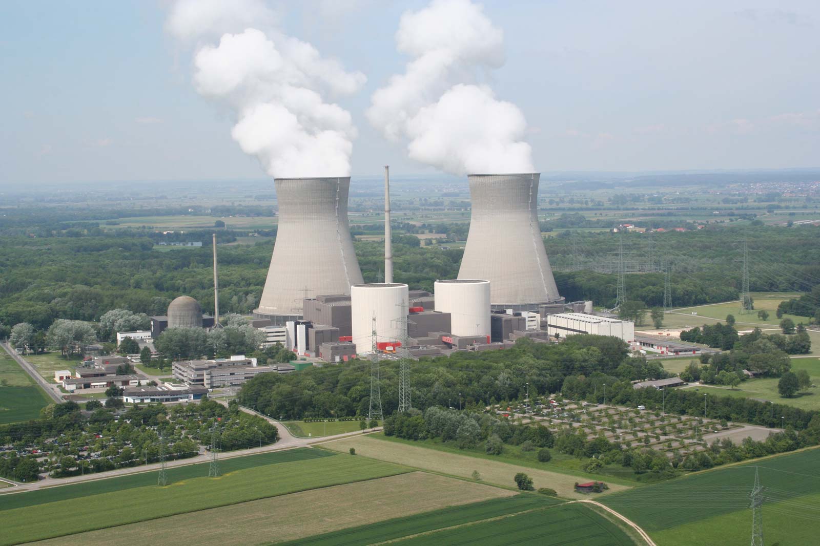 A nuclear power plant featuring cooling towers, steam clouds, and surrounding fields and trees.