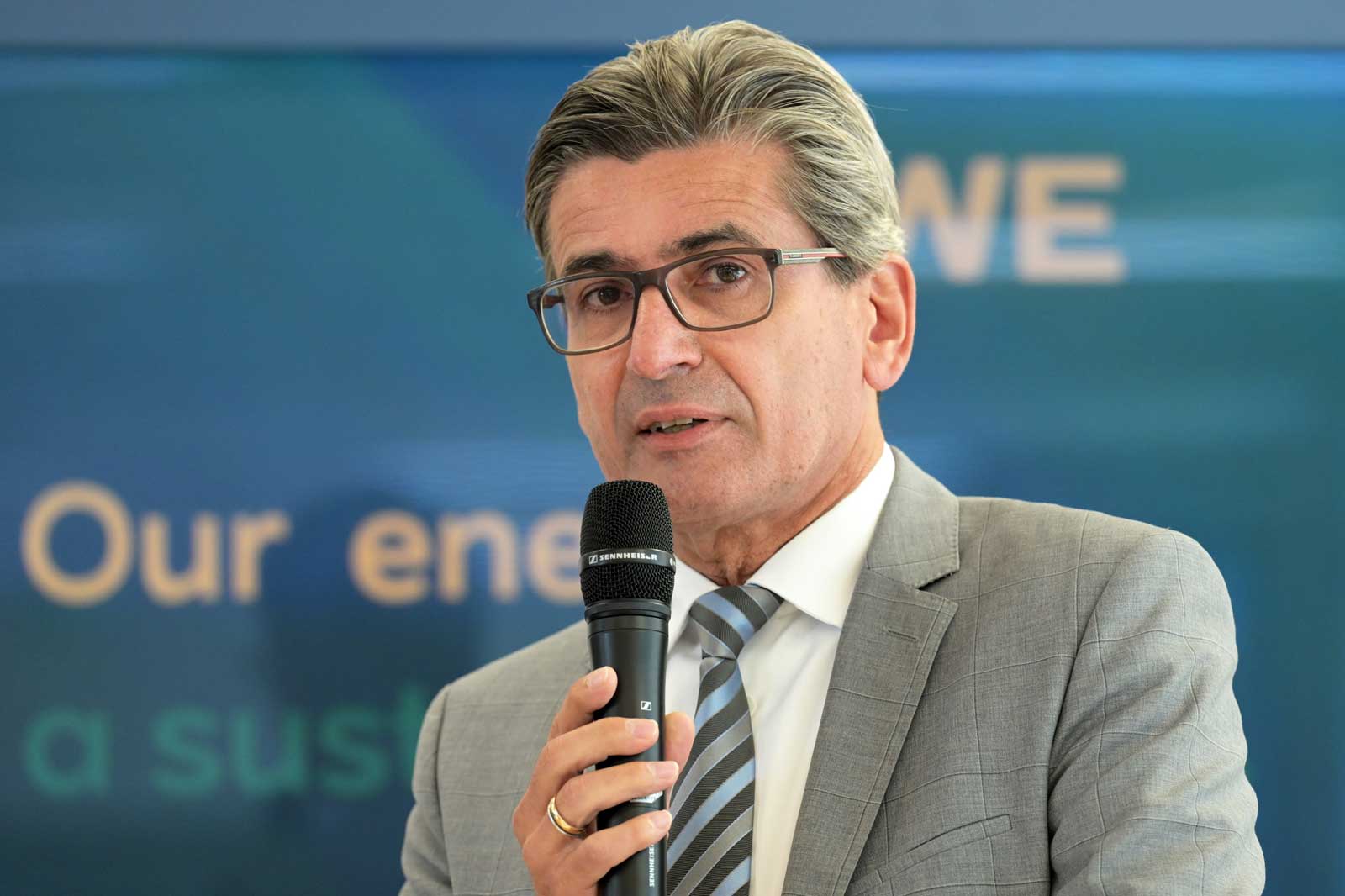 Ein Redner in einem grauen Anzug hält ein Mikrofon und spricht vor einem blauen Hintergrund mit Text.