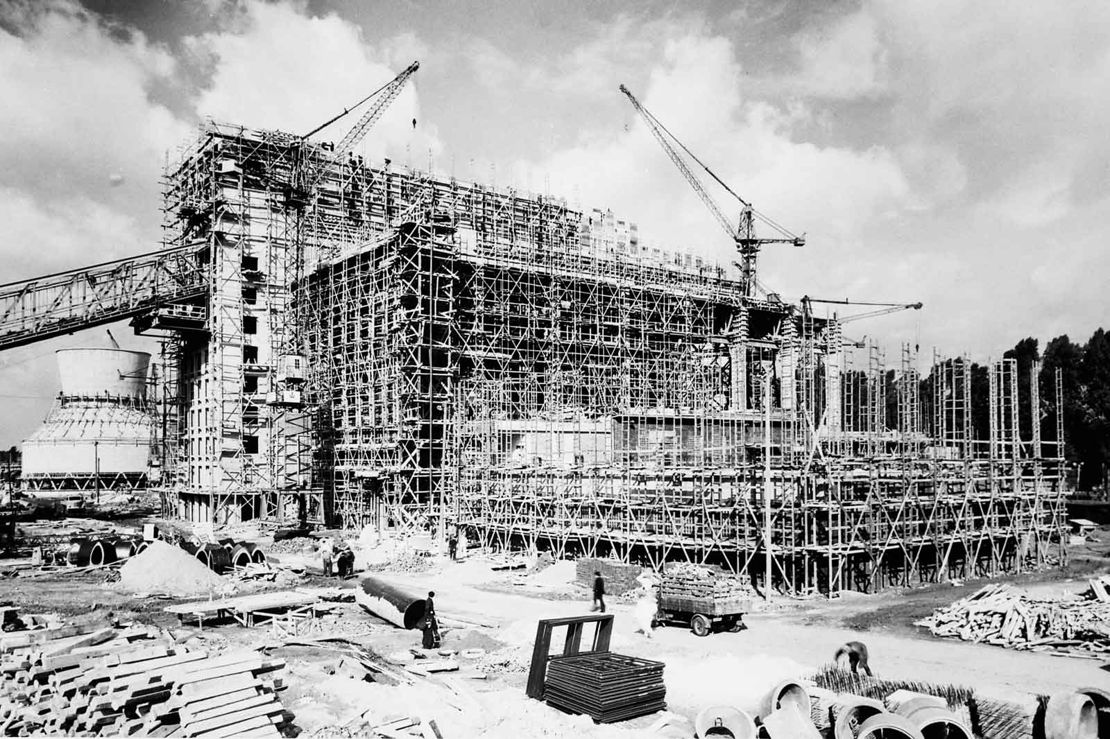 Eine große Baustelle mit Kränen und Gerüsten. Arbeiter sind sichtbar, und ein Kühlturm ist im Hintergrund.