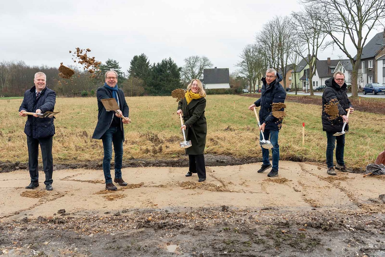 Fünf Personen nehmen an einer Grundsteinlegung teil, während sie Schaufeln halten und Erde in einem Grasbereich werfen.