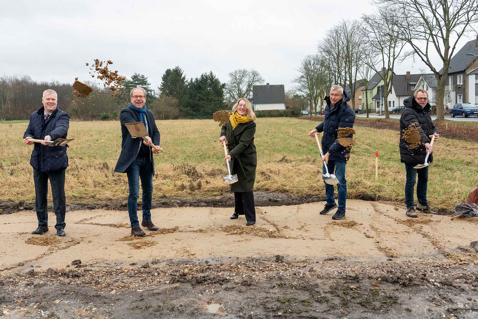 Fünf Personen nehmen an einer Grundsteinlegung teil, während sie Schaufeln halten und Erde in einem Grasbereich werfen.