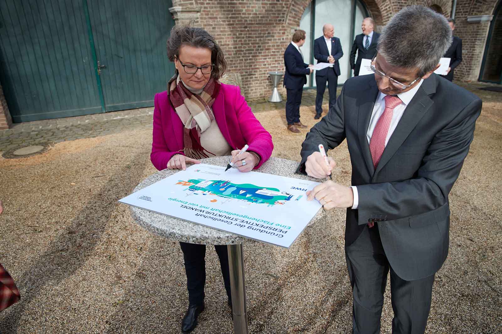 Zwei Personen unterschreiben ein großes Dokument auf einem Tisch, während mehrere Personen im Hintergrund stehen. Die Szene ist im Freien.