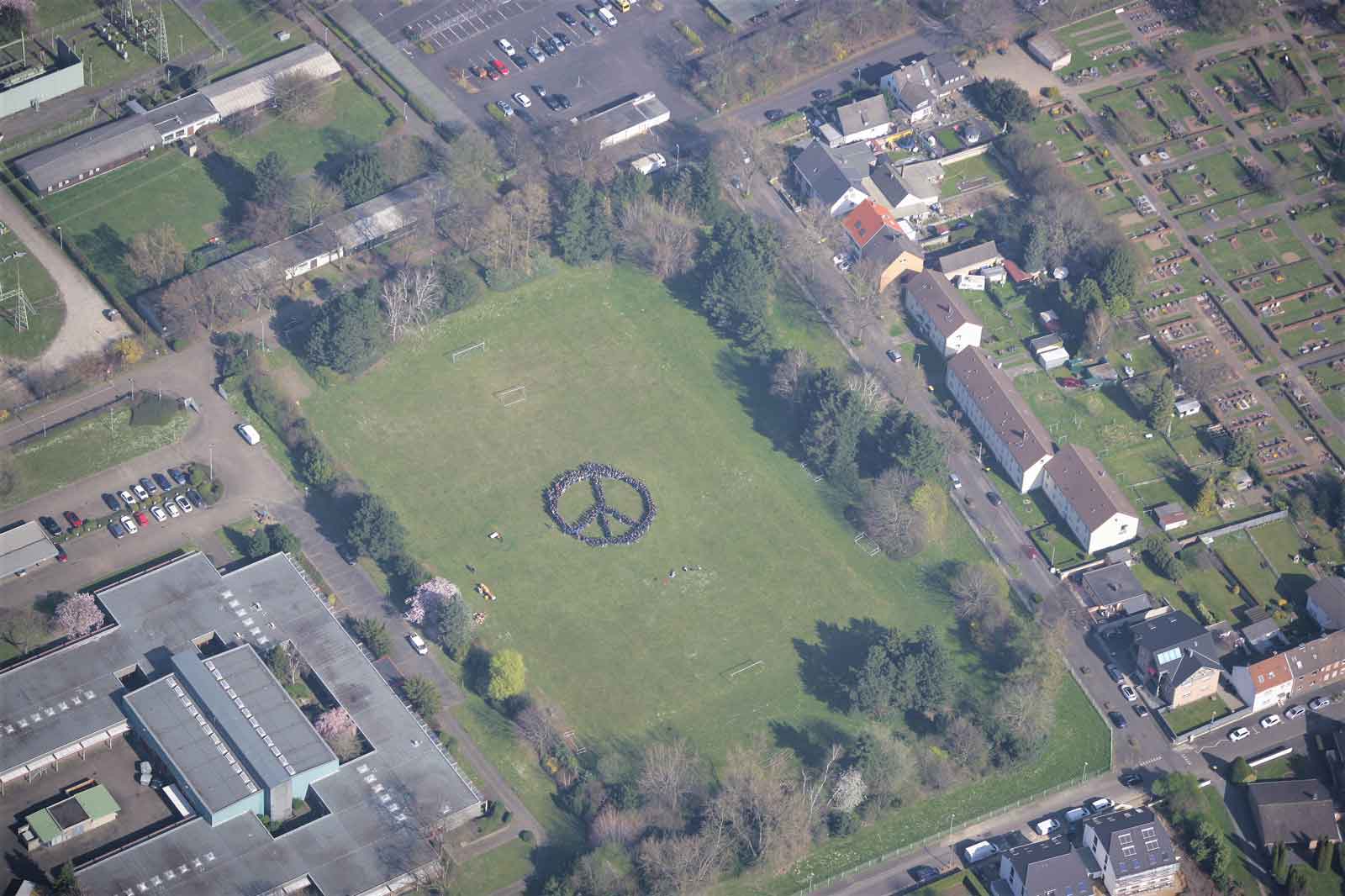 Eine Luftaufnahme eines Parks mit einem großen Frieden-Symbol, das von Menschen gebildet wird, umgeben von Häusern und Bäumen.