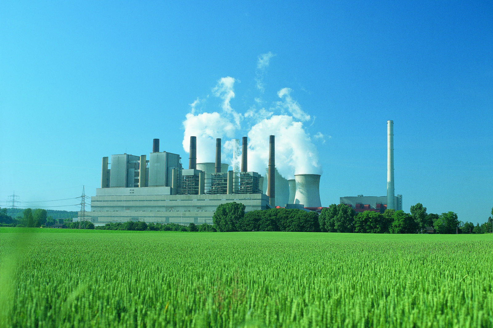 Ein Kraftwerk umgeben von grünen Feldern unter einem klaren blauen Himmel, mit Rauch, der aus Kühltürmen aufsteigt.