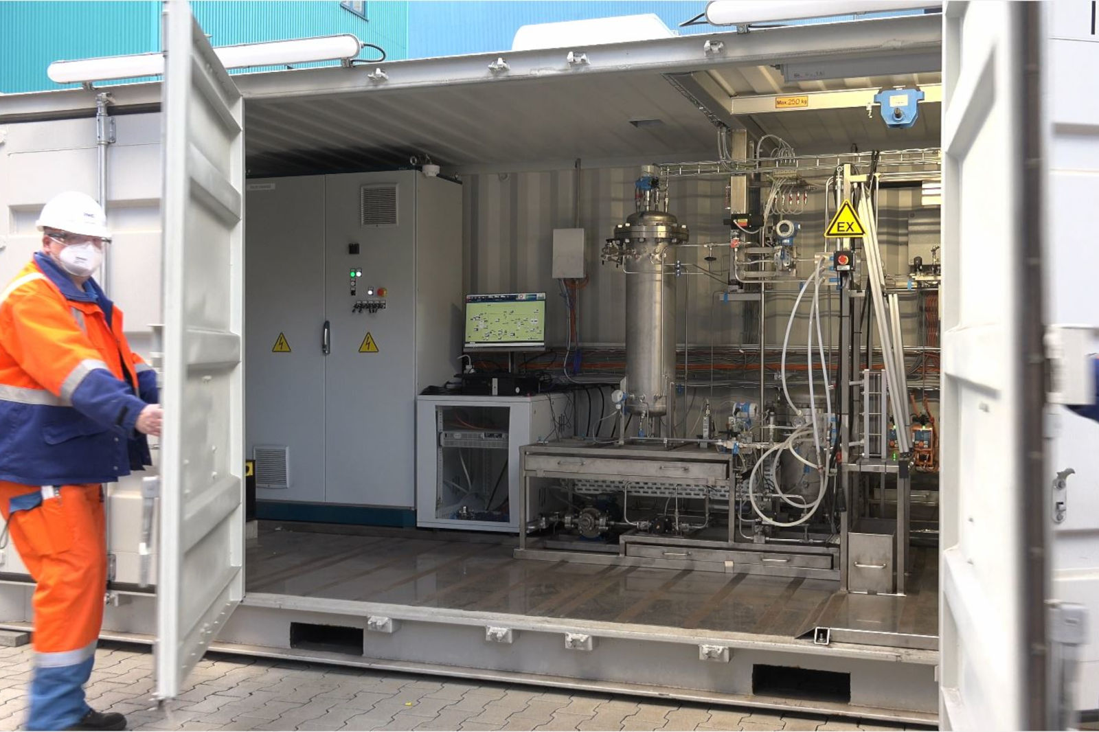 Ein Techniker in orange-blauer Uniform steht am Eingang eines Versandcontainers und zeigt die darin enthaltene Industrieausrüstung.