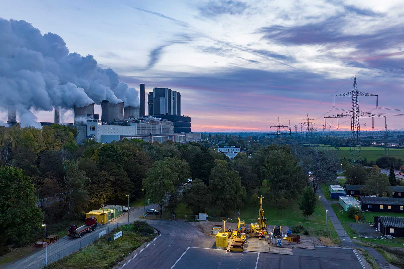 Ein Kraftwerk, das Rauch ausstößt, mit Strommasten und grünen Feldern unter einem Dämmerungshimmel.