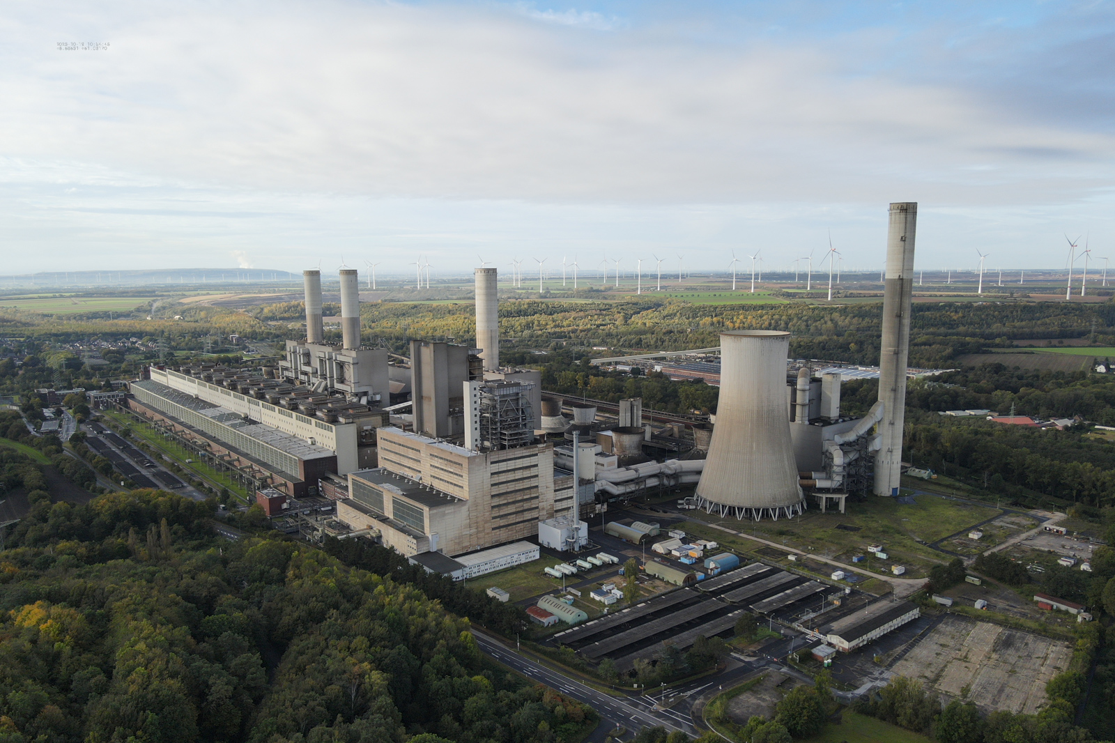 Eine Luftaufnahme eines Industriekraftwerks mit Schornsteinen und Windkraftanlagen im Hintergrund, umgeben von grünem Landschaft.