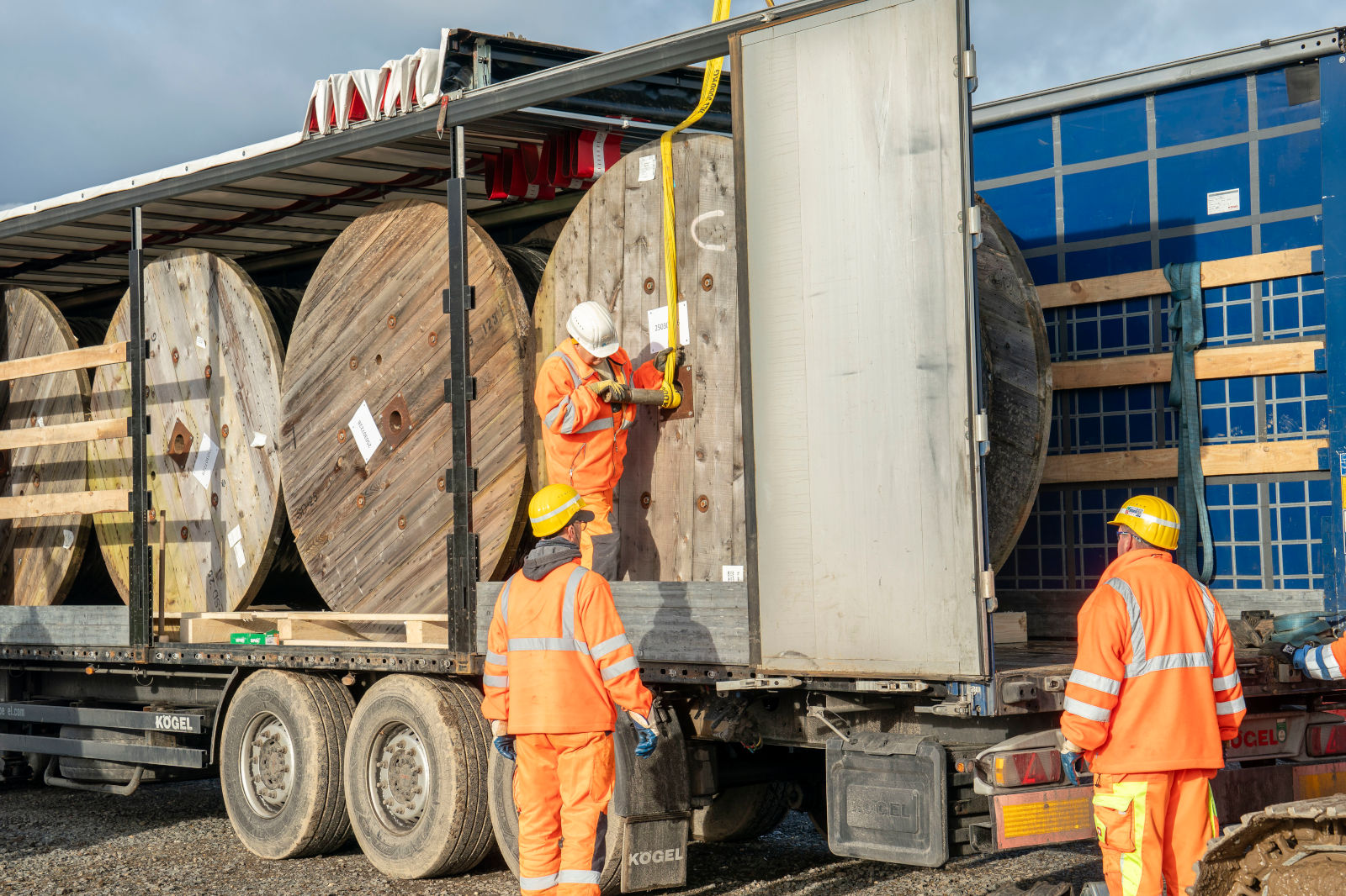 Arbeiter in orangefarbener Sicherheitskleidung laden Holzspulen auf einen Lkw und sichern sie mit Gurten und Equipment.