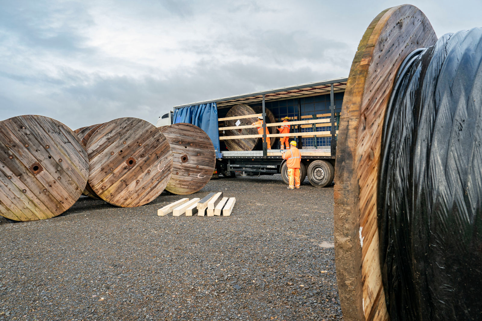 Zwei Arbeiter in orangenen Overalls laden große Holzspulen aus einem Lkw unter einem wolkigen Himmel aus.
