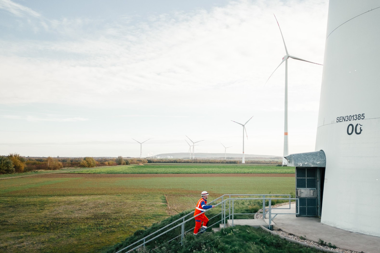 Ein Arbeiter in einem roten Overall steigt eine Treppe neben einer Windkraftanlage in einem grünen Feld mit anderen Turbinen im Hintergrund hinauf.