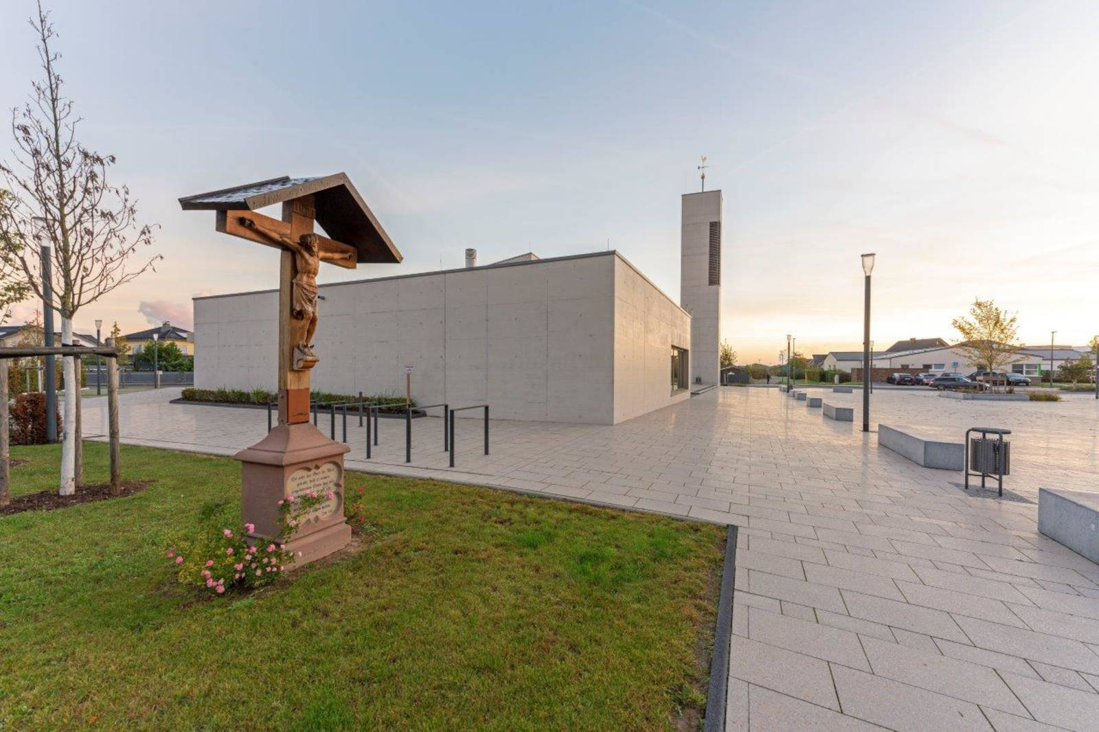 Ein modernes Gebäude mit einem hohen Glockenturm. Ein hölzernes Kreuz steht davor, umgeben von Gras und Blumen.
