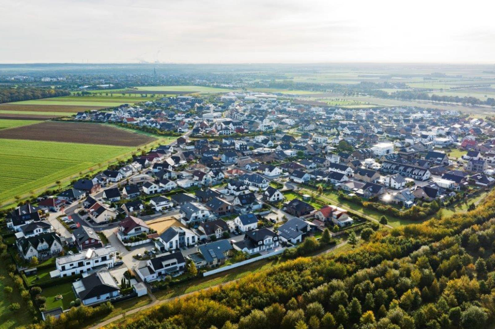 Luftaufnahme eines Vororts mit Wohnhäusern, grünen Feldern und Bäumen unter einem bewölkten Himmel.
