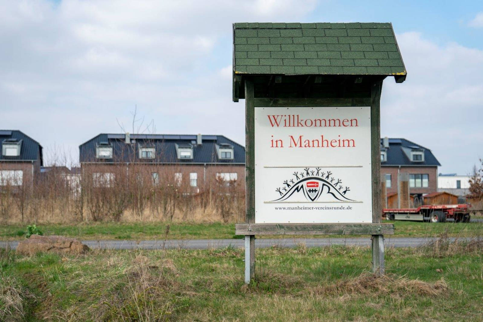 Willkommensschild in Mannheim mit grünem Dach, zeigt lokale Website, umgeben von Wohngebäuden und Gras.