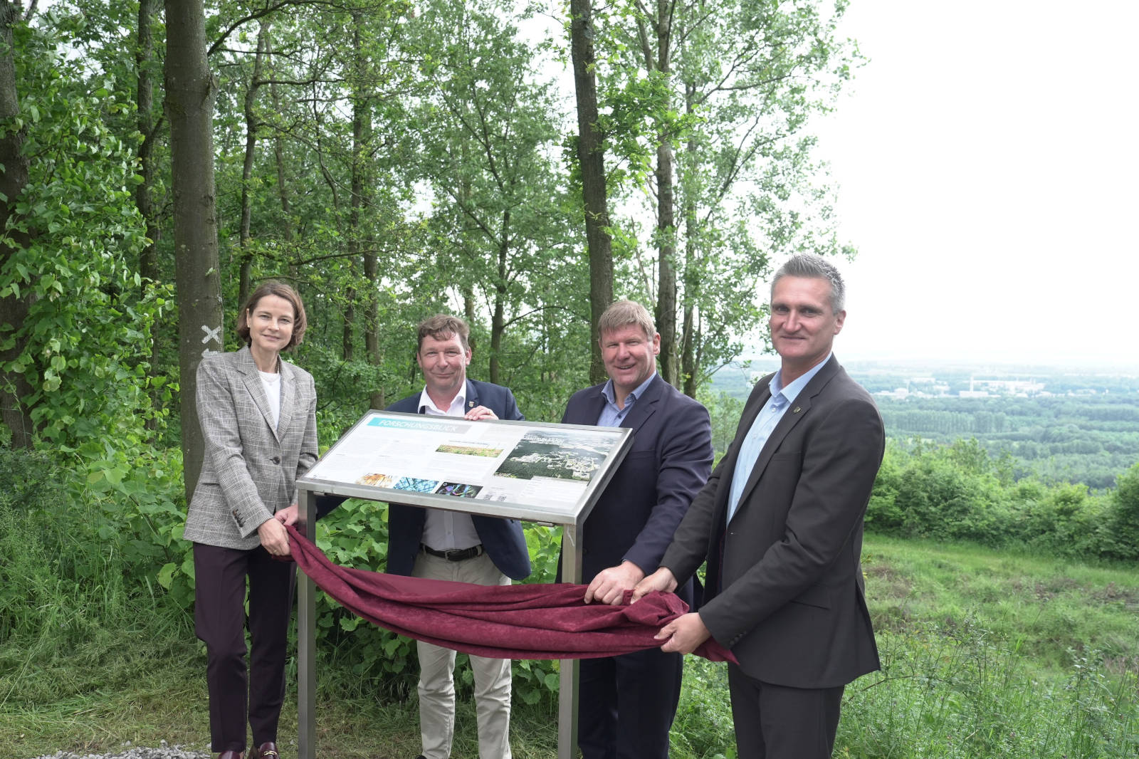 Vier Personen enthüllen ein Infoboard in einem üppigen grünen Wald mit einer fernen Aussicht.
