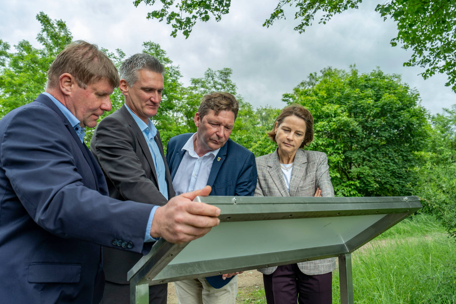 Eine Gruppe von vier Personen steht um ein Informationsschild in einer grünen Außenumgebung und führt eine Diskussion.