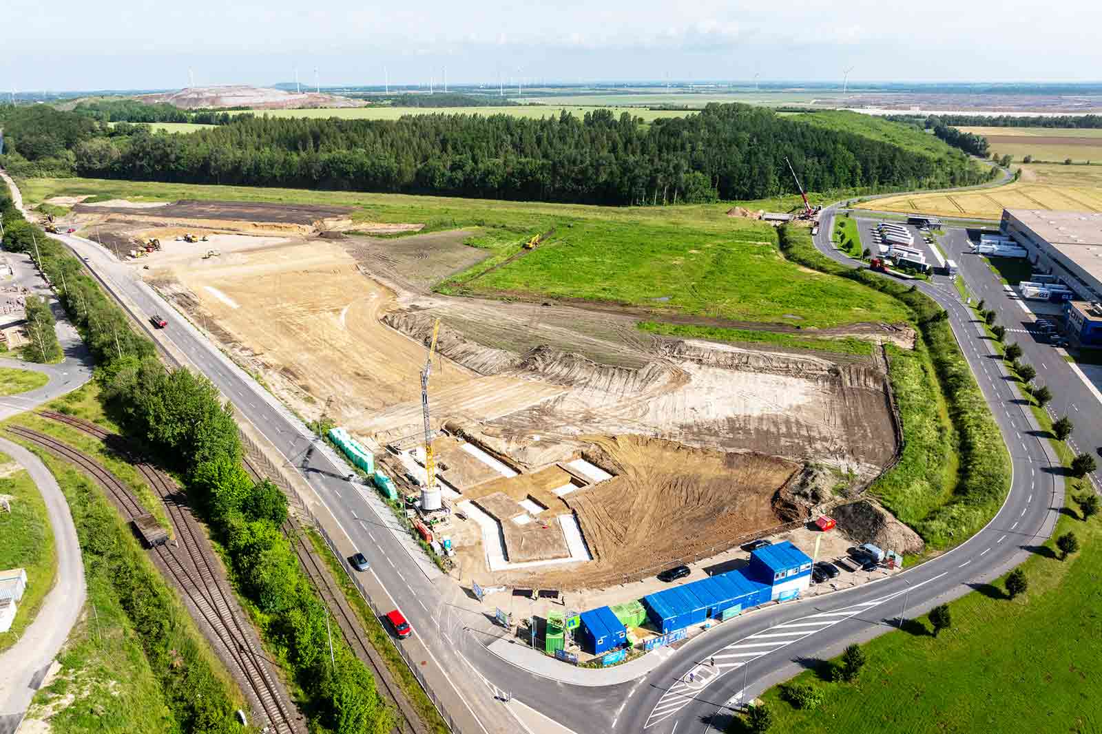Eine Luftaufnahme einer Baustelle mit Maschinen, geräumtem Gelände und nahegelegenen Straßen inmitten grüner Felder.