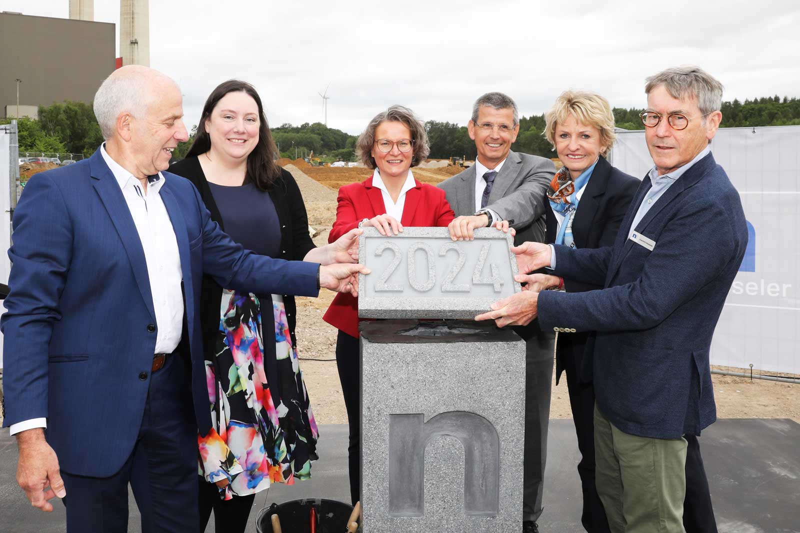 Gruppe von Personen, die an einer Grundsteinlegung mit einem Stein mit der Aufschrift '2024' an einer Baustelle teilnehmen.