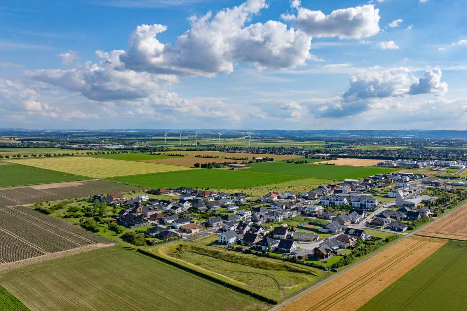 Luftaufnahme eines Dorfes, umgeben von grünen Feldern, mit blauem Himmel und Wolken darüber.