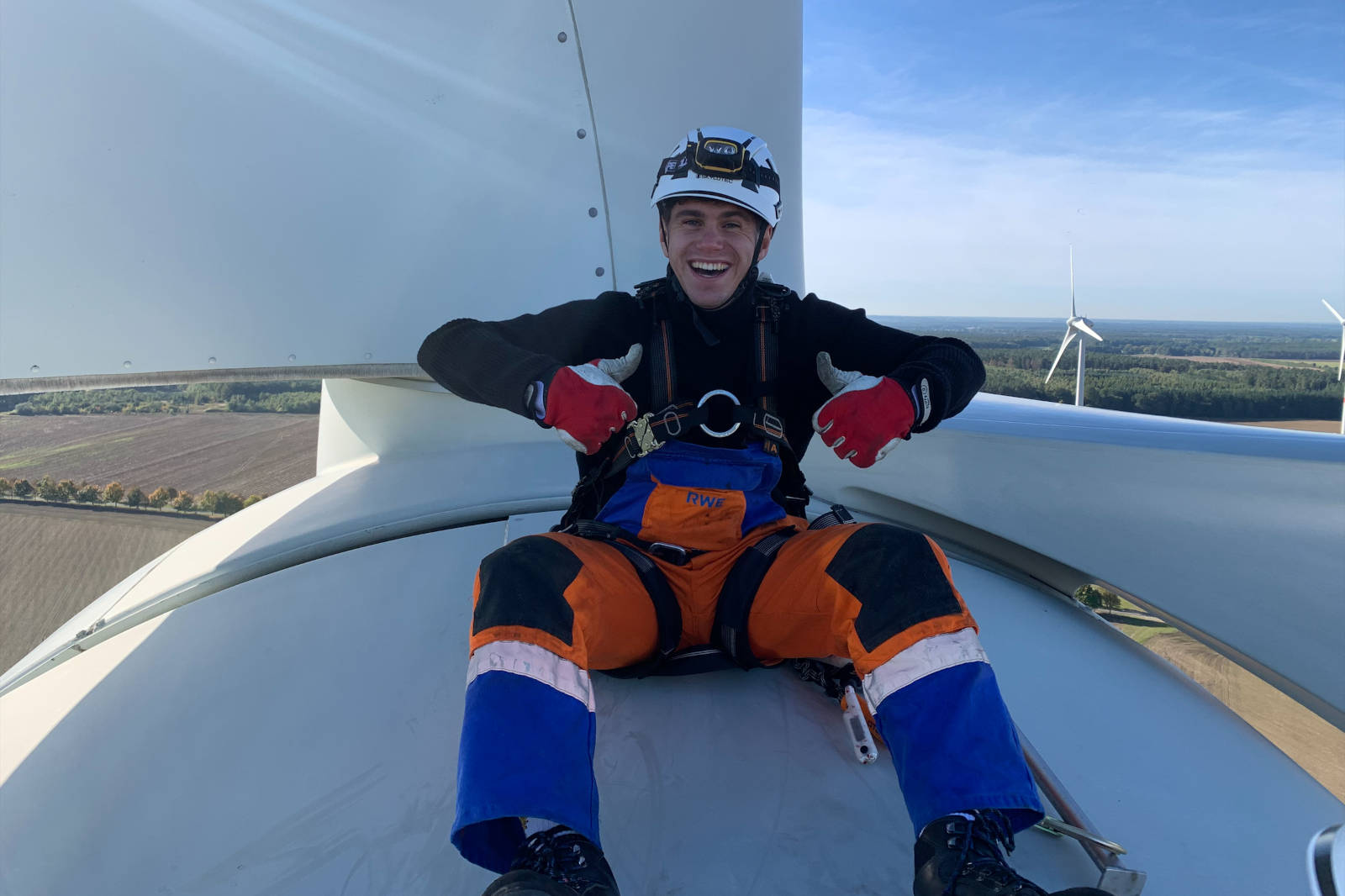 Ein Arbeiter sitzt auf einem Windrad, trägt Sicherheitsausrüstung und zeigt den Daumen nach oben, mit Blick auf Felder und Windräder in der Ferne.