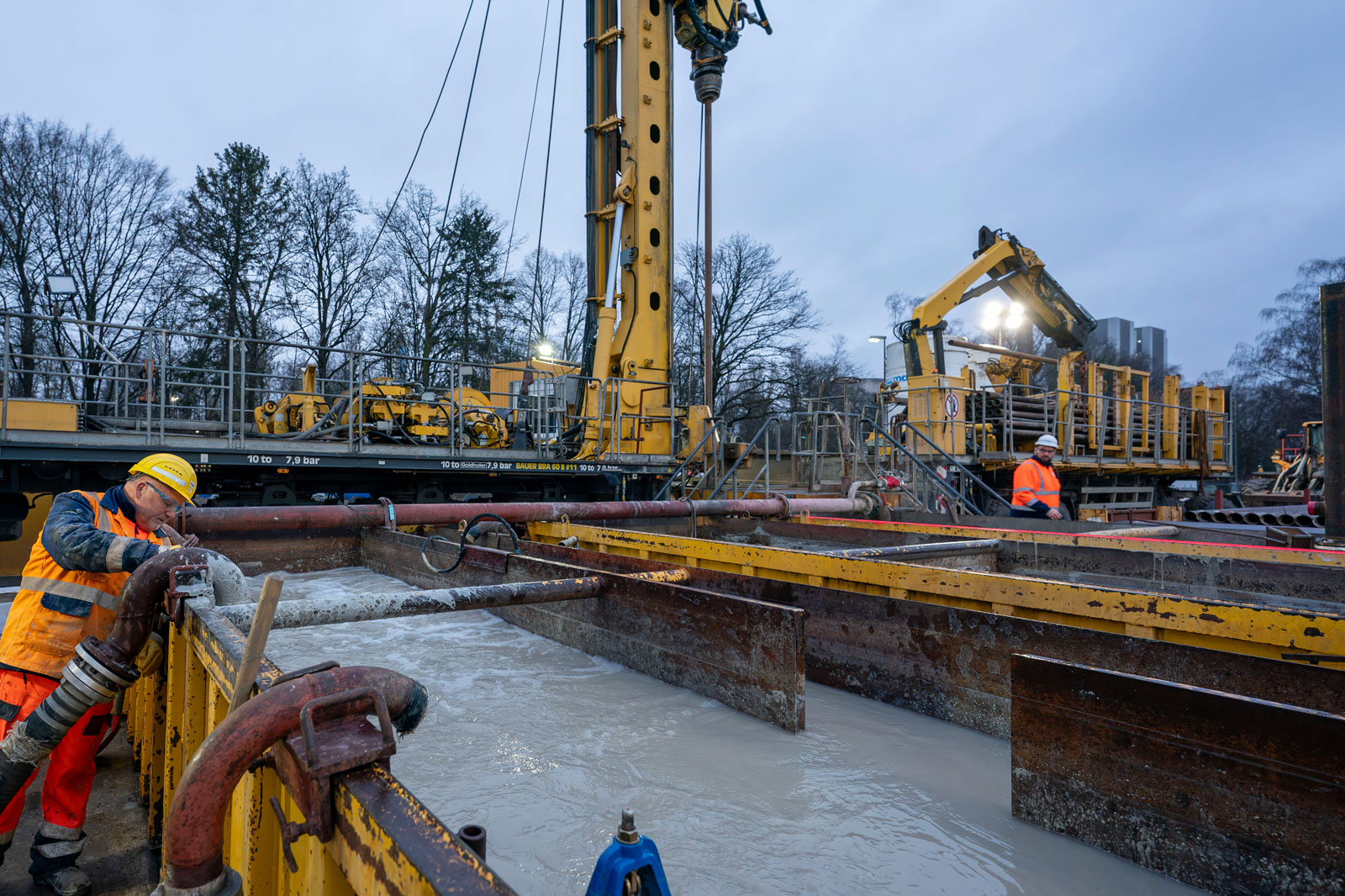 Bauarbeiter bedienen Geräte an einer großen Baustelle mit Maschinen und Wassertanks unter einem bewölkten Himmel.