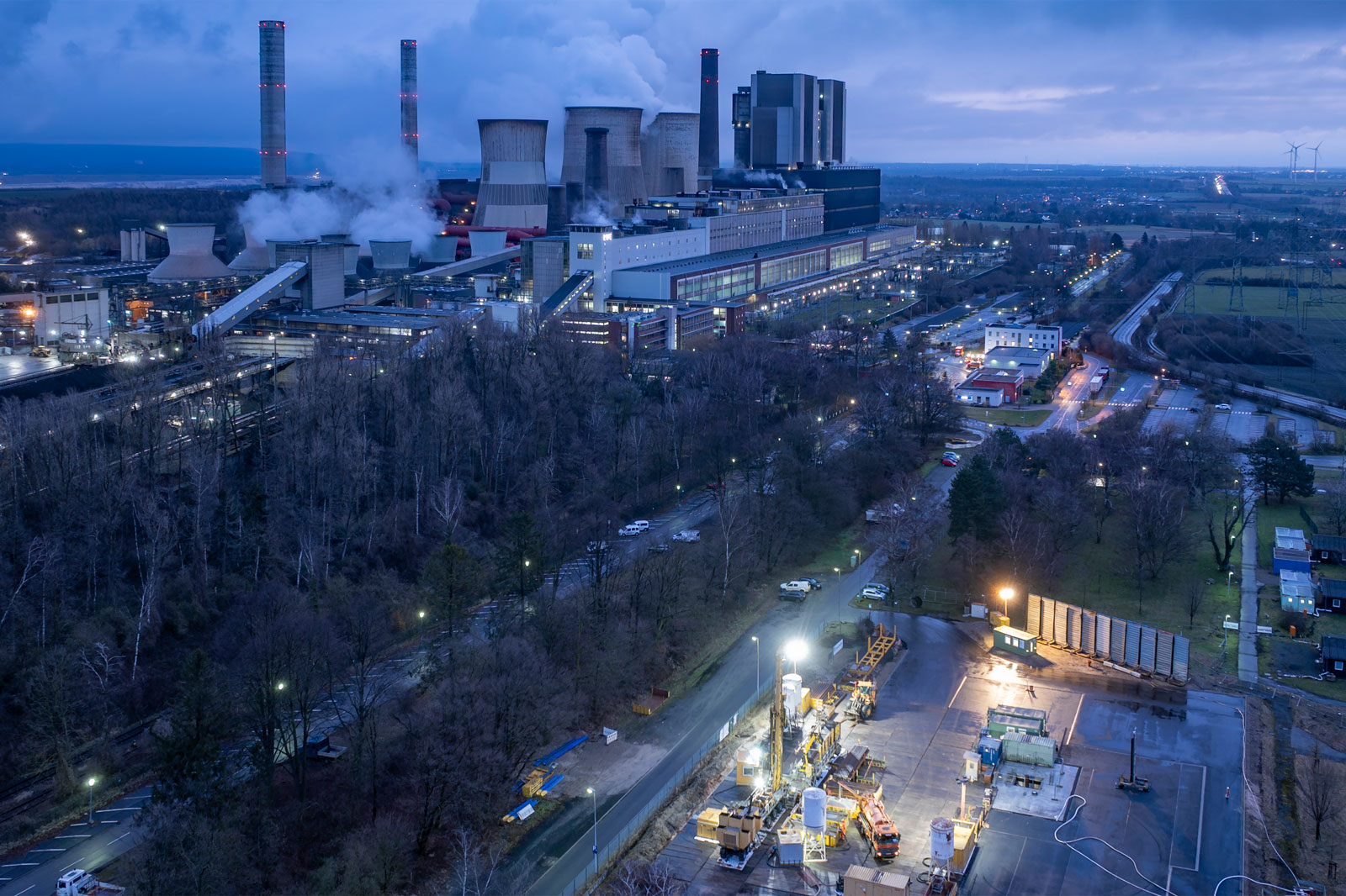 Ein Kraftwerk mit Schornsteinen und Kühltürmen, bei Dämmerung beleuchtet, neben Straßen und Bäumen.