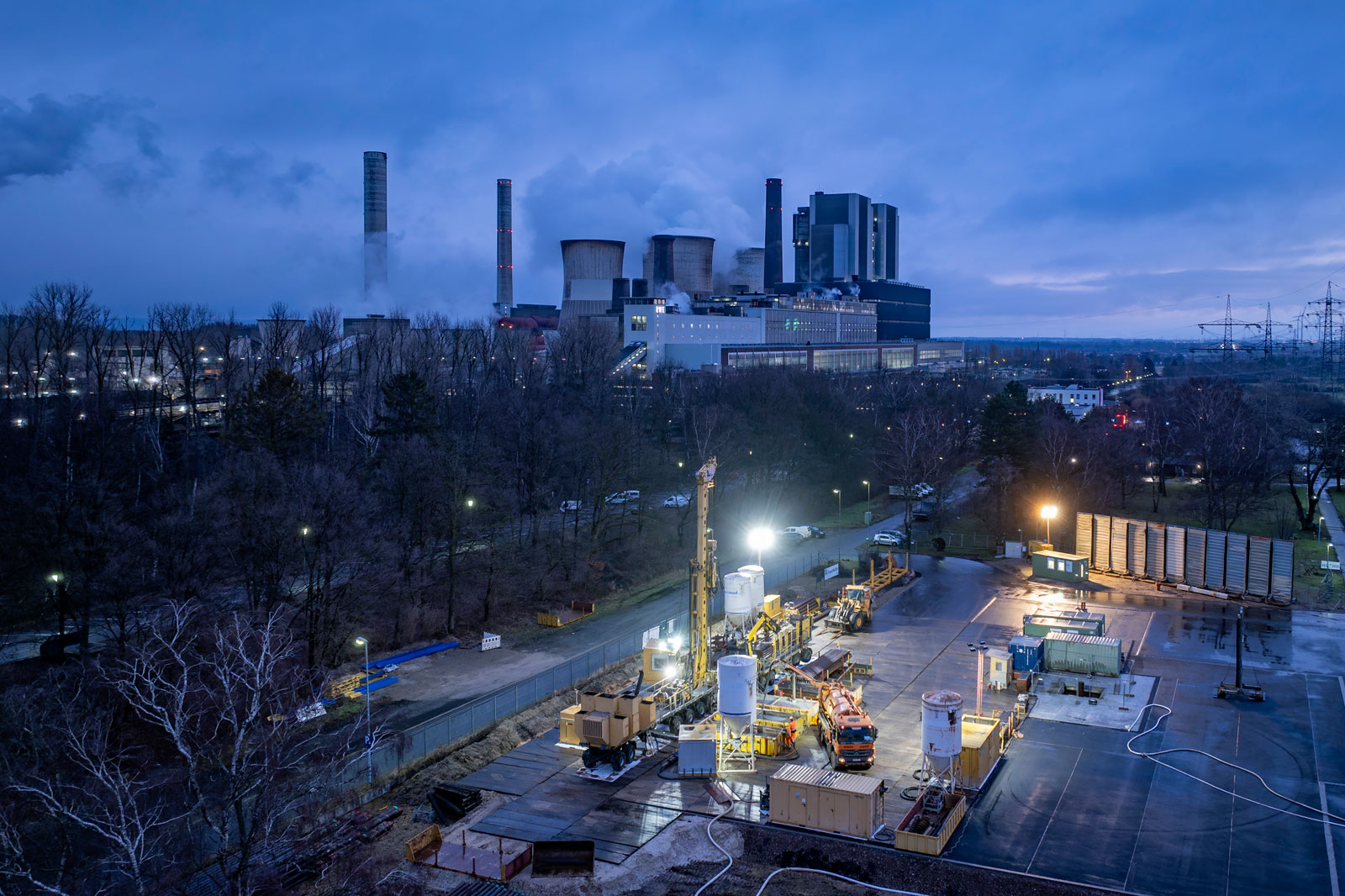 Eine Aussicht auf ein Kraftwerk bei Dämmerung, mit Kühltürmen und Industrieanlagen, die von Kunstlicht beleuchtet sind.
