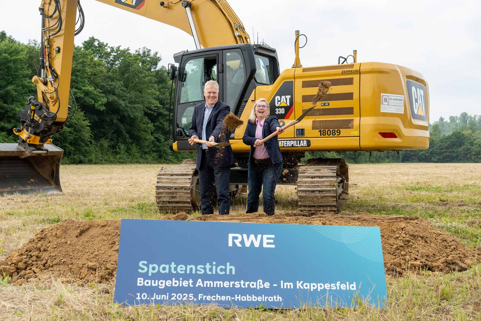 Zwei Männer in Anzügen halten Schaufeln an einer Baustelle mit einem großen Bagger im Hintergrund und einem Schild, das das Projekt ankündigt.