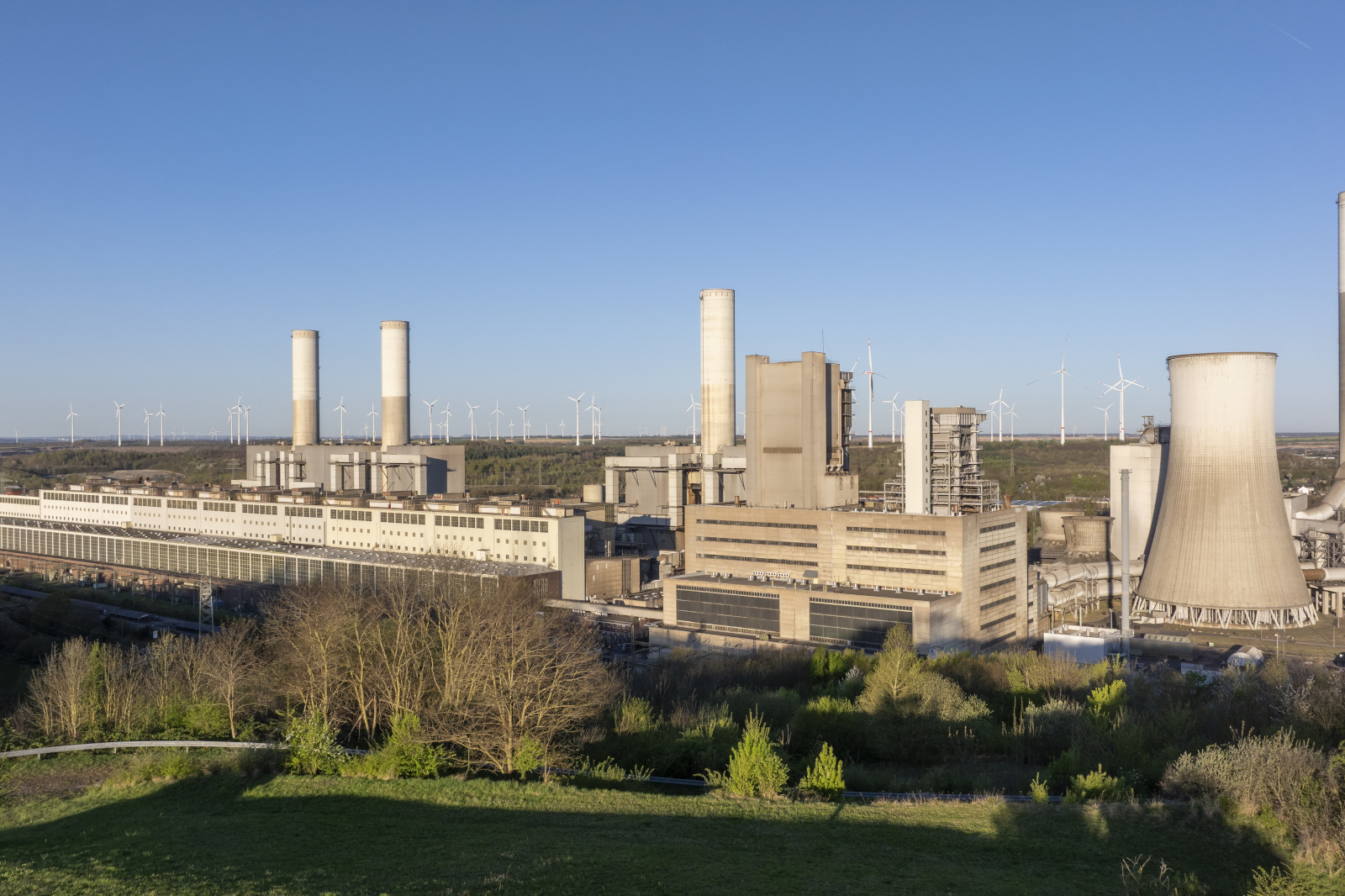 Ein Kraftwerk mit Kühltürmen und Schornsteinen, umgeben von grünen Feldern und Windkraftanlagen unter einem klaren blauen Himmel.