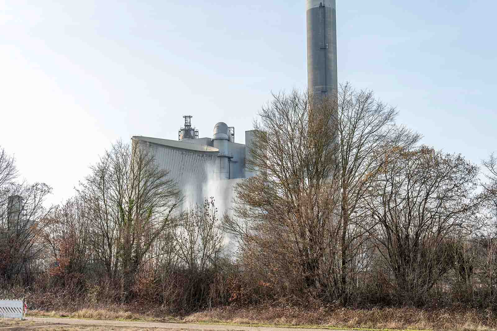 Große Industrieanlage mit Silos und hohem Schornstein, aus dem Dampf steigt, vor kahlen Bäumen; links ist eine weiße Absperrung sichtbar.