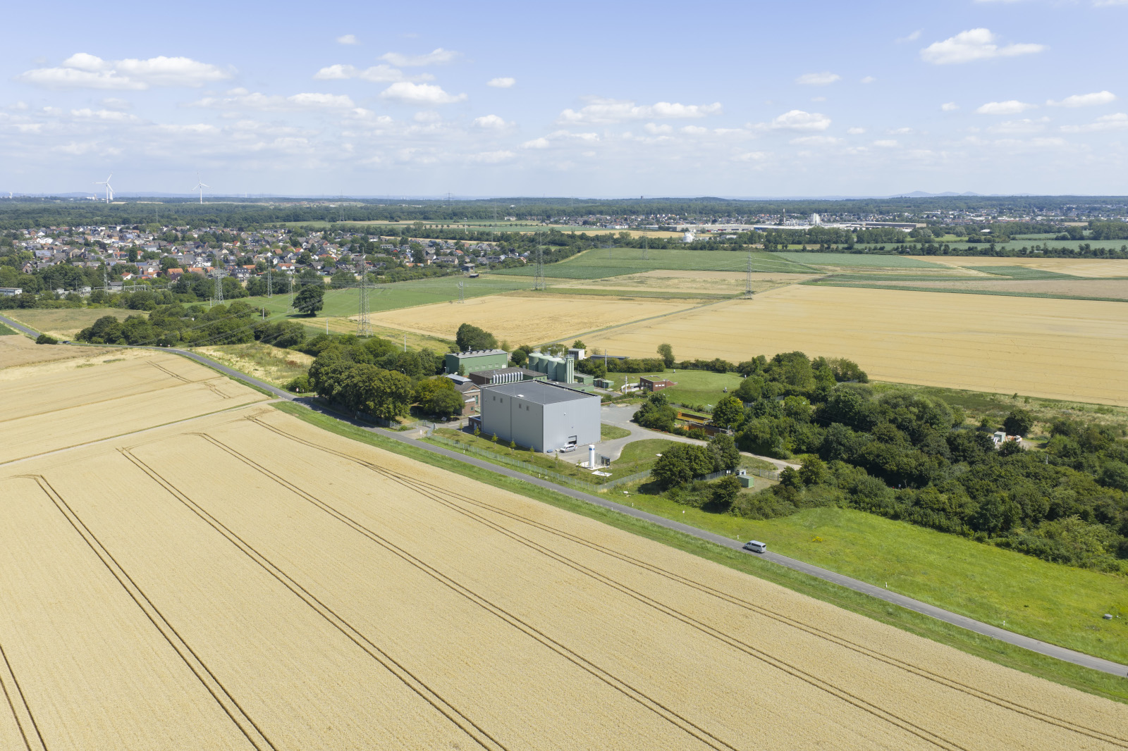 Im Vordergrund weite goldene Getreidefelder, in der Bildmitte ein graues Industriegebäude aus Metall und Beton neben grünen Bäumen und einer Landstraße unter blauem Himmel.