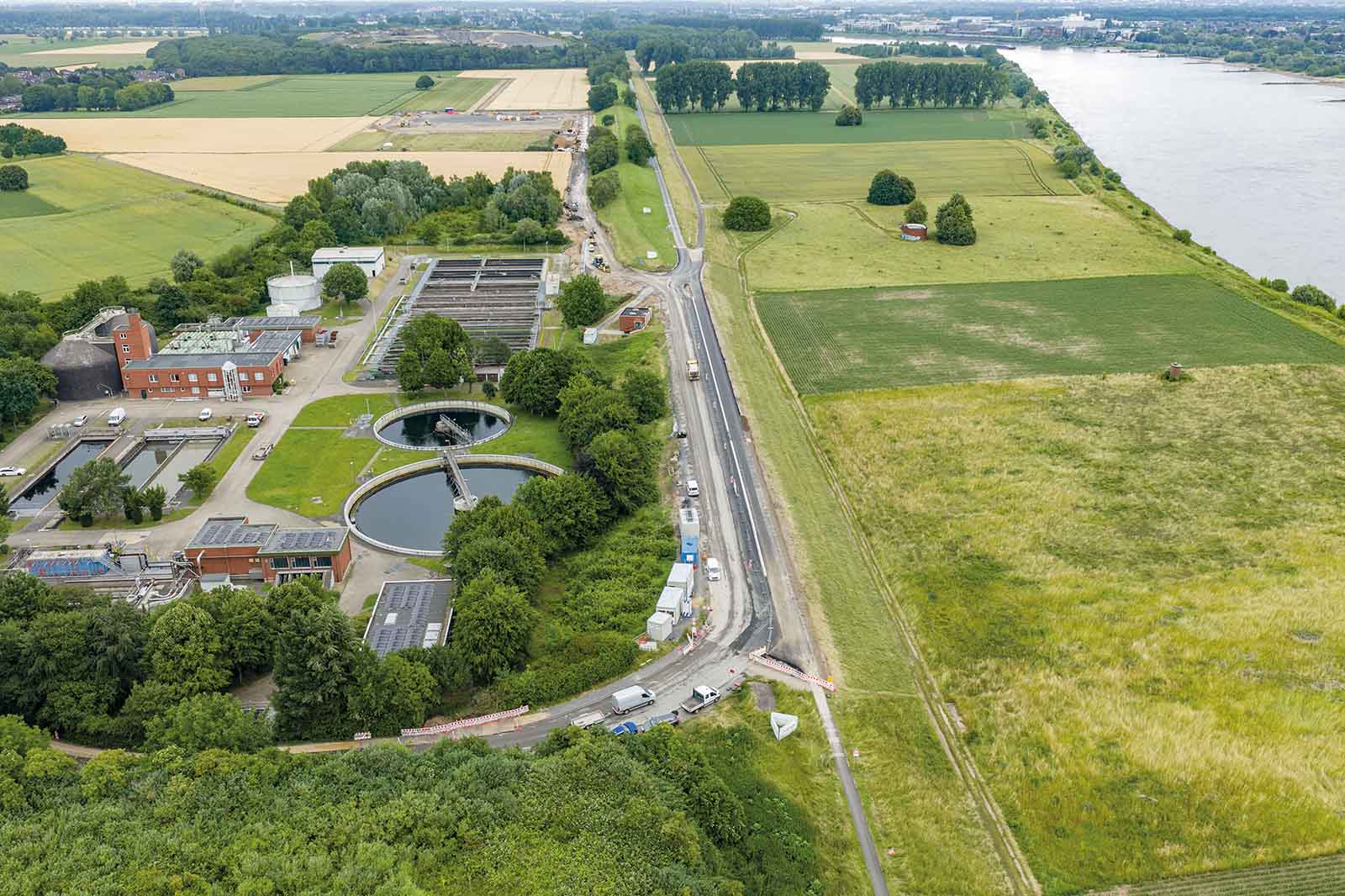 Im Vordergrund eine Kläranlage mit runden Betonbecken neben einer asphaltierten Landstraße; dahinter grüne Felder und ein breiter Fluss unter hellem Himmel.