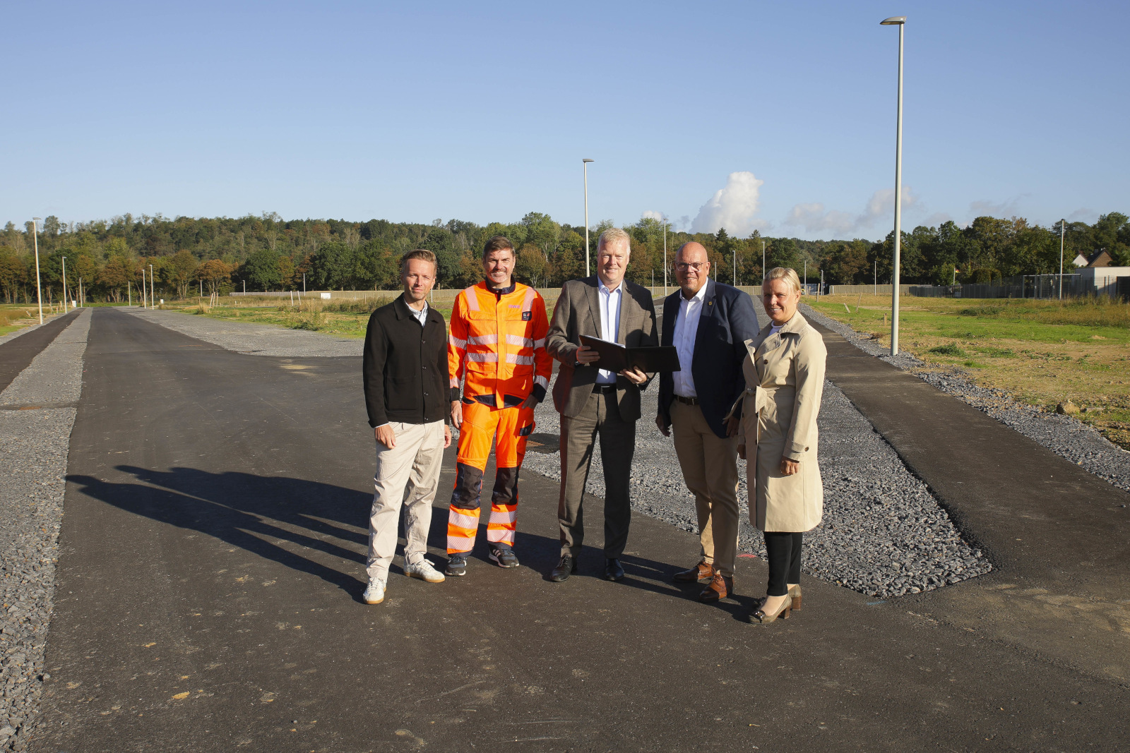 Fünf Personen stehen auf einer frisch asphaltierten Straße vor Bäumen und blauem Himmel; ein Mann trägt orange Warnkleidung, andere Anzug und Trenchcoat.