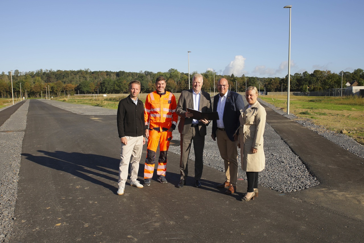 Fünf Personen stehen auf einer frisch asphaltierten Straße vor Bäumen und blauem Himmel; ein Mann trägt orange Warnkleidung, andere Anzug und Trenchcoat.
