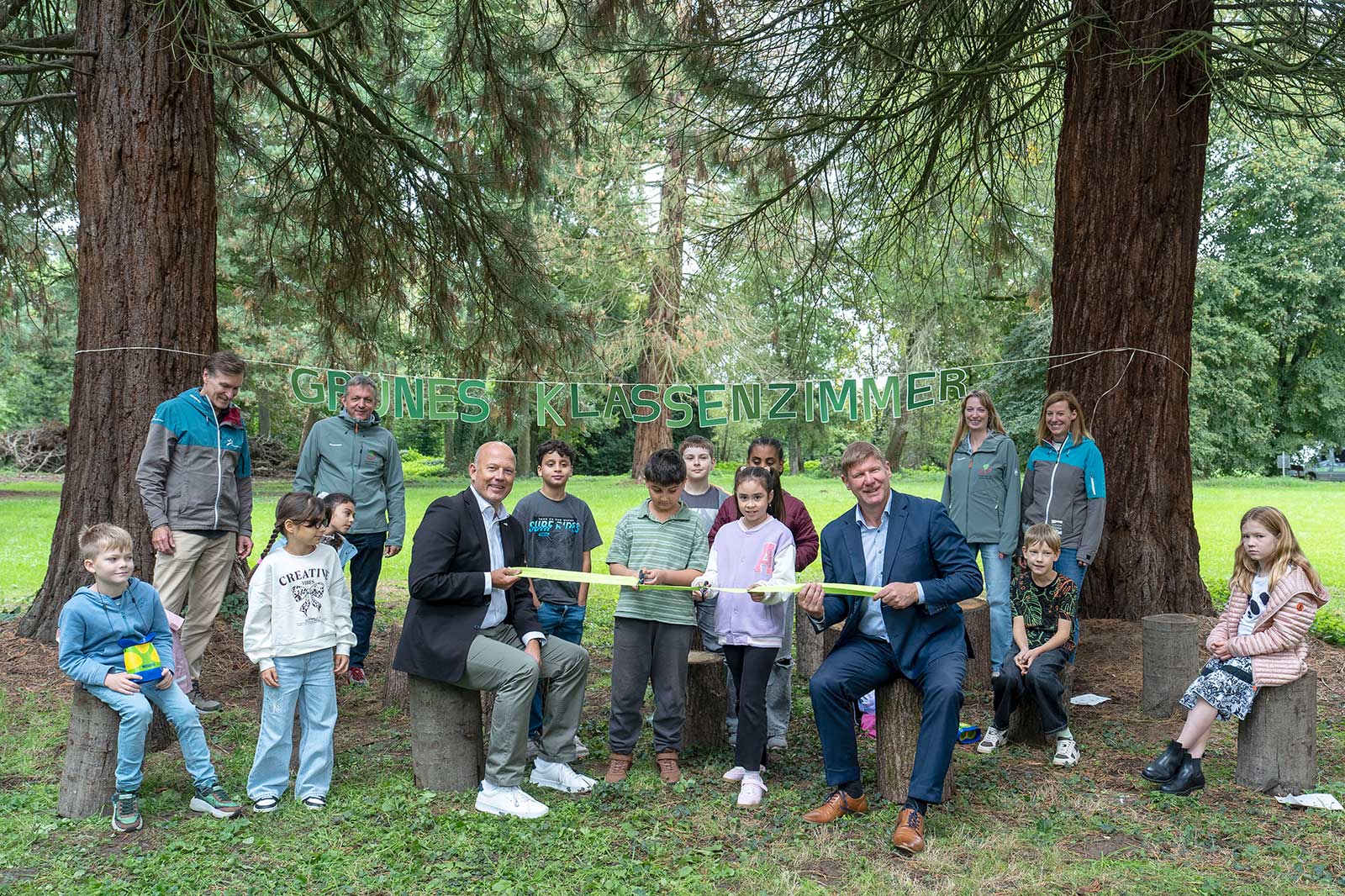 Zwei Männer in Anzügen halten vorne ein grünes Band, umringt von lächelnden Kindern und Erwachsenen auf Baumstümpfen. Im Hintergrund hängen zwischen hohen Bäumen grüne Buchstaben mit der Aufschrift „GRÜNES KLASSENZIMMER“.