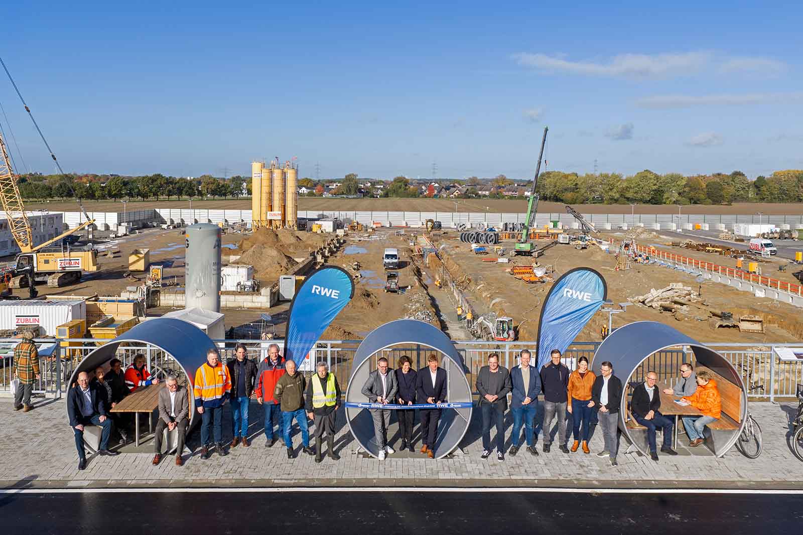 Personengruppe in Anzügen und orangenen Sicherheitswesten steht in großen runden Betonrohren auf einer Baustelle mit Kränen und zwei blauen Fahnen „RWE”.