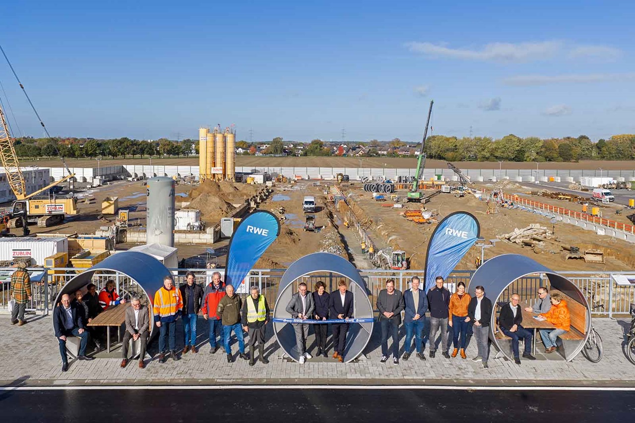Personengruppe in Anzügen und orangenen Sicherheitswesten steht in großen runden Betonrohren auf einer Baustelle mit Kränen und zwei blauen Fahnen „RWE”.