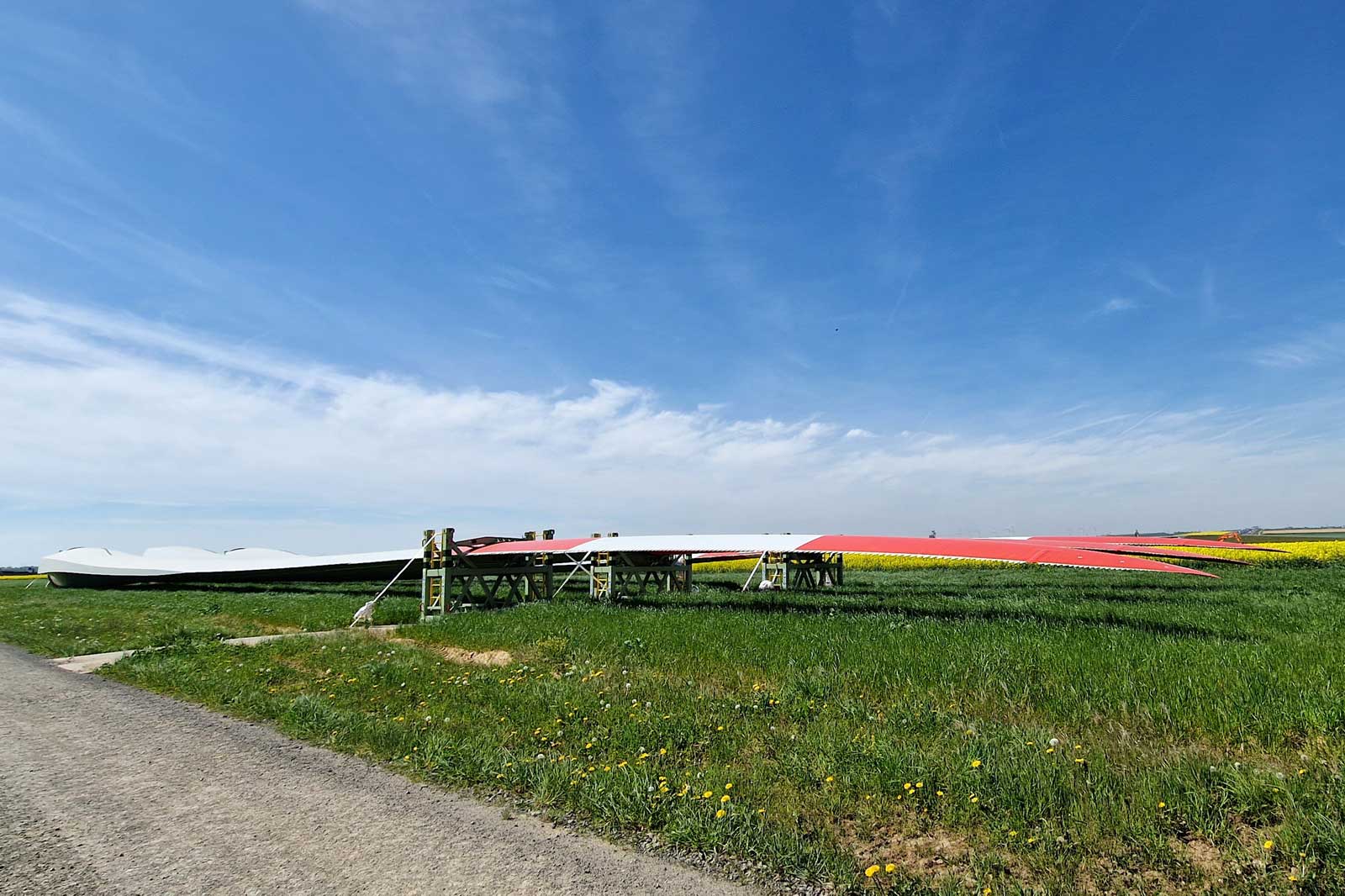 Große Windturbinenschaufeln liegen auf Stützen in einem grünen Feld unter einem blauen Himmel mit vereinzelten Wolken.