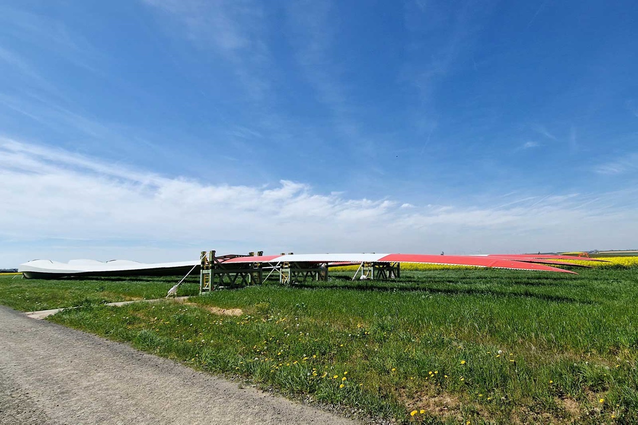 Large wind turbine blades lie on supports in a green field under a blue sky with scattered clouds.