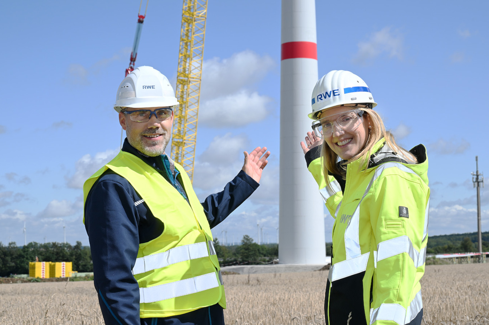 Two people in work clothing stand in front of a wind turbine, surrounded by a field landscape.