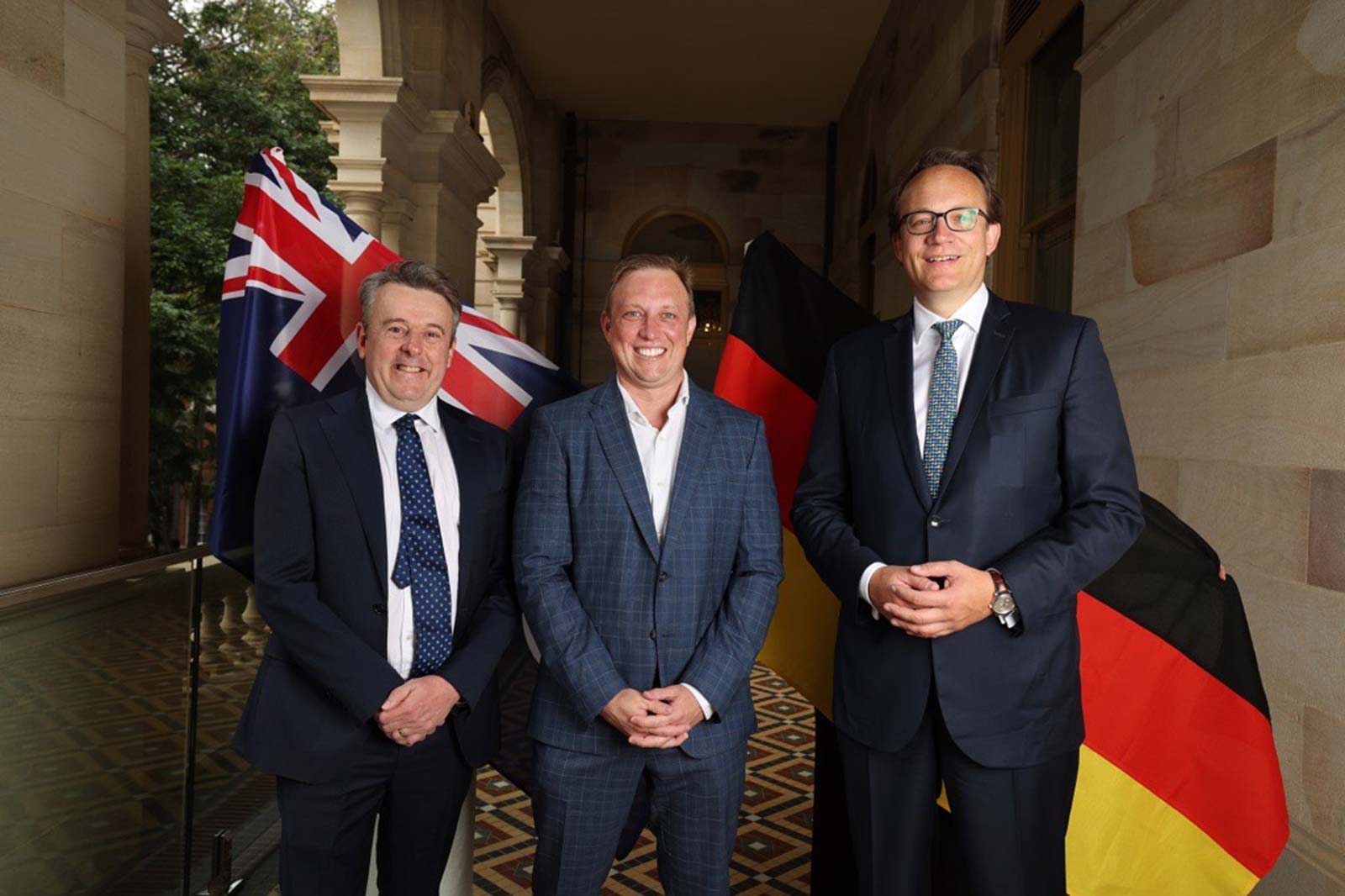 Three individuals pose with Australian and German flags in a historic building setting. They are dressed in formal attire.