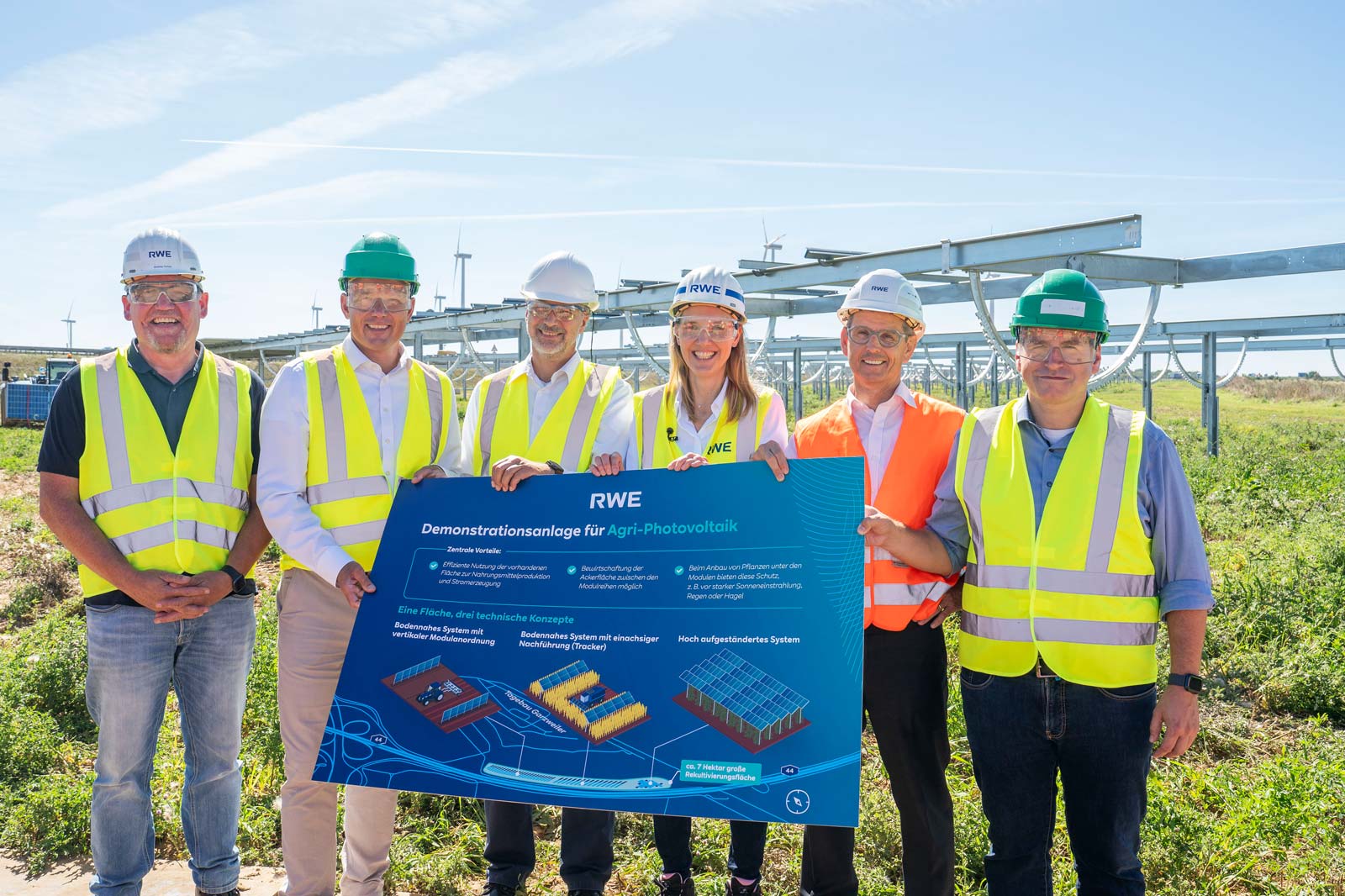 Five individuals in safety vests and helmets present an information poster about Agri-Photovoltaics in a sunny field.
